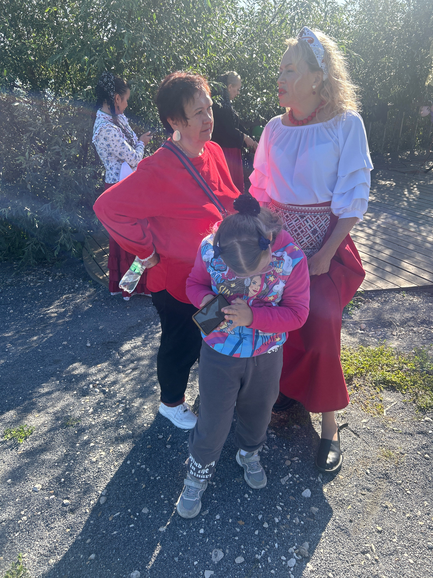 In an outdoor setting, three individuals are captured. The foreground features a young girl looking down at a smartphone in her hands, wearing a vibrant pink and blue jacket with cartoon graphics and grey pants. To her left, an adult woman in a bright red long-sleeved shirt and black pants converses with another adult. The second adult, standing to the girl's right, is a woman with blonde hair wearing a white blouse with ruffled sleeves, a red beaded necklace, and a long red skirt with a decorative embroidered band. In the background, another woman in a floral shirt and a woman in a black jacket are visible, with lush green foliage forming the backdrop. The ground consists of gravel with patches of grass and dappled sunlight. The scene appears to be a casual gathering in an outdoor public space.