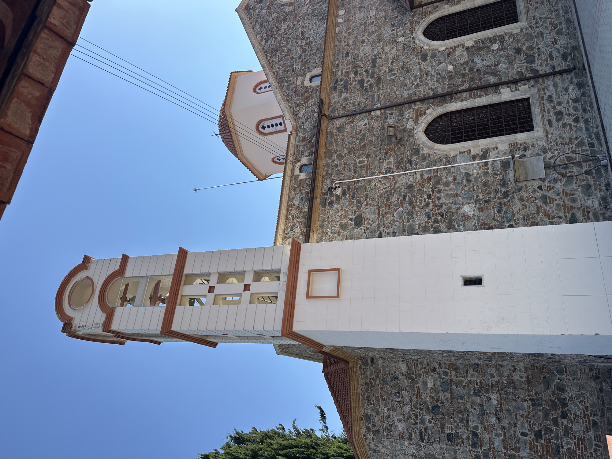 The image shows the exterior of a stone church with a white bell tower. The bell tower is tall and has several arched openings with bells visible. It is decorated with orange trim. The church itself is made of rough-hewn stones and has a red-tiled roof. There are arched windows with metal grates on the side of the church. A dome, also with a red-tiled roof and arched windows, is visible on top of the church. The sky is clear and blue. A tall green tree is visible to the left of the bell tower. The scene is captured from a low angle, looking up towards the structures. The overall impression is of an old, traditional religious building in a sunny environment. The church is identified as the Church of Panagia Eleousa in Agros, Cyprus.