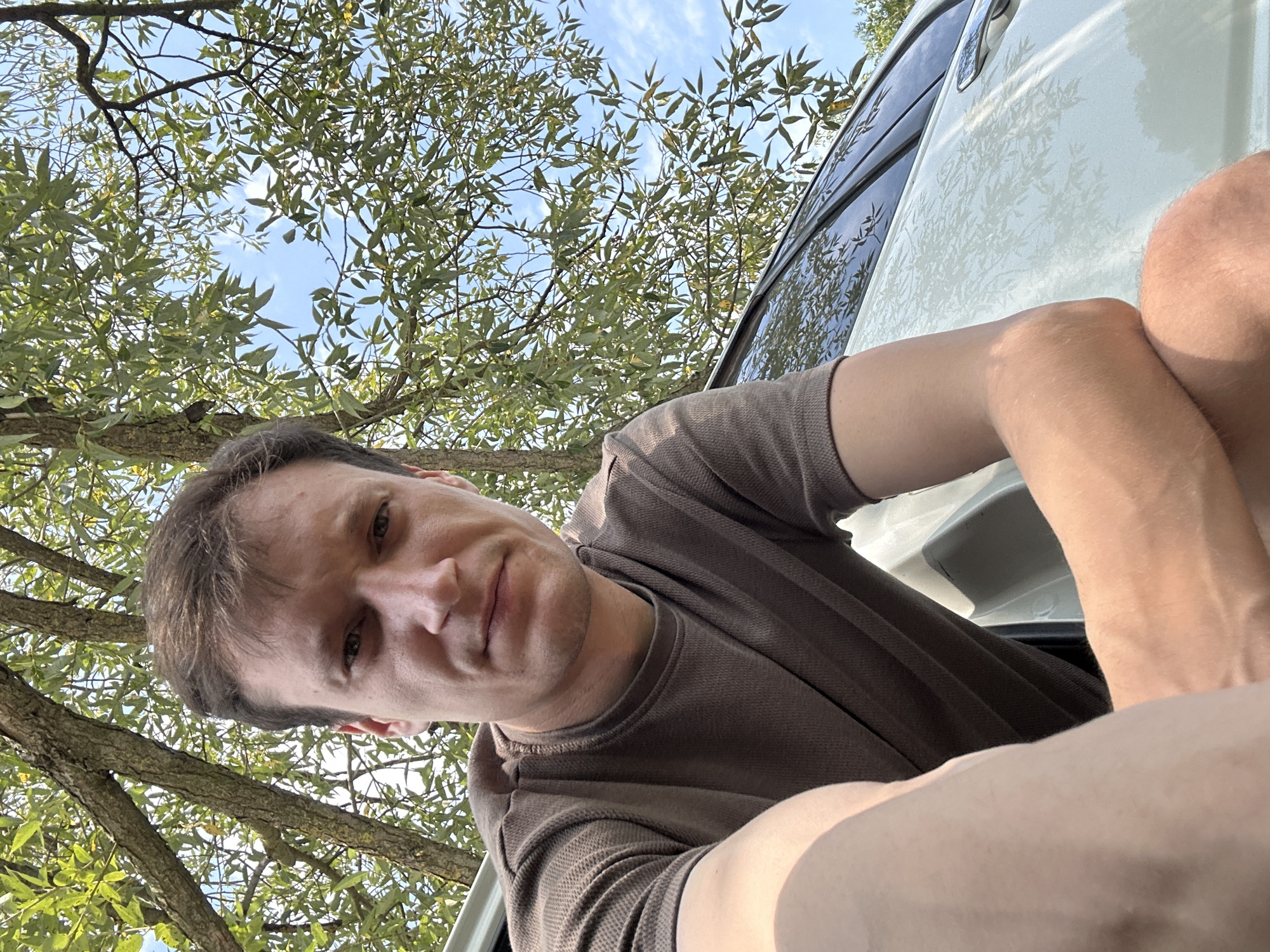 A man with brown hair and a brown t-shirt is captured in a selfie, with his face angled towards the camera. He is outdoors, with a white car visible to his right and a tree with green leaves and branches above him. The sky is blue and clear. The image is taken at eye-level, with the man's arms resting on his knees. The background suggests a natural or park-like setting, potentially in or near Makiv, Ukraine.