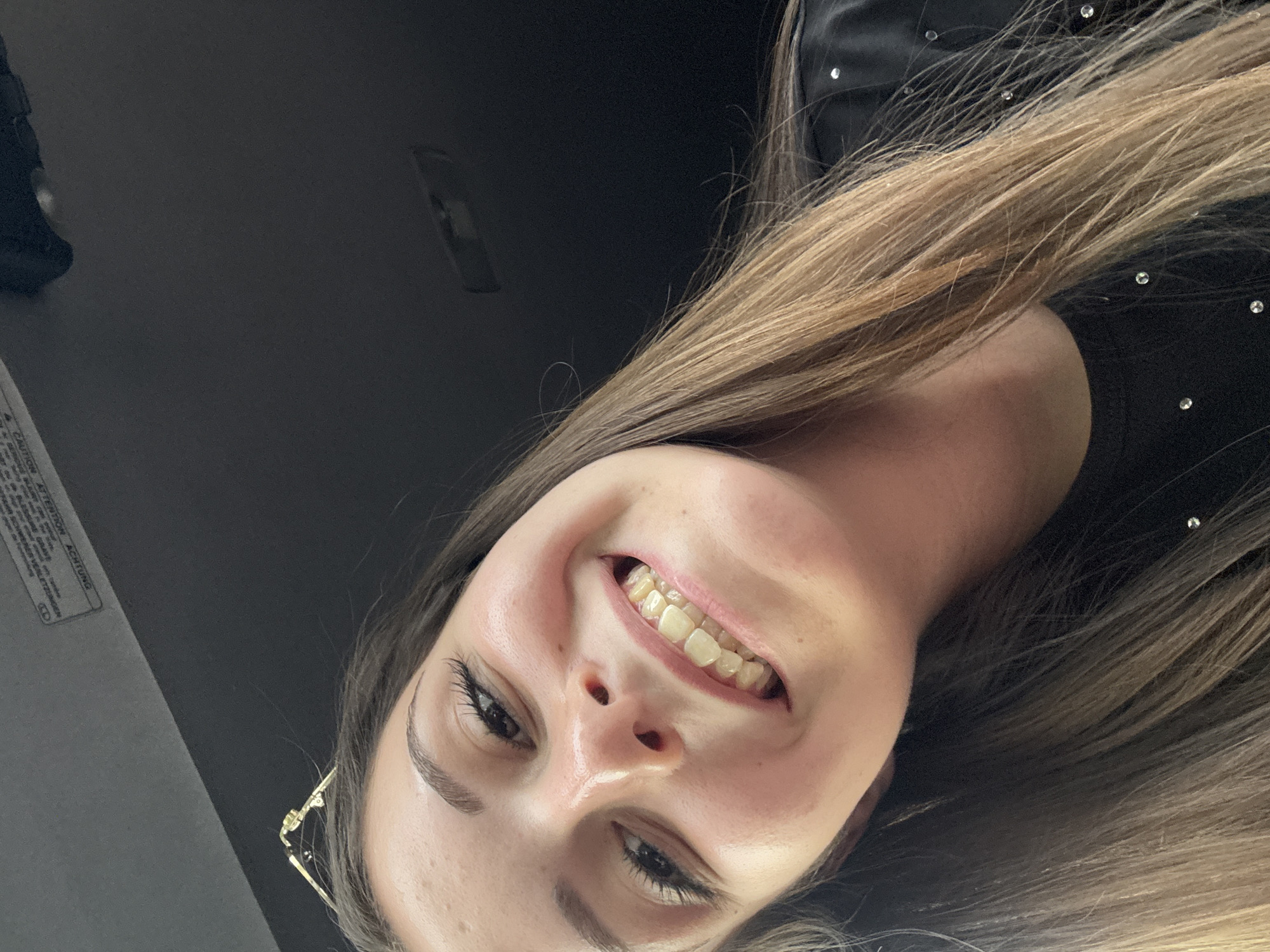 A young woman with brown hair and fair skin is taking a selfie. She is smiling, showing her teeth which appear to be yellow. She is wearing a black shirt with rhinestones. The background is dark, suggesting an indoor environment. There is a label visible at the top of the image with German text, possibly from a car's sun visor. The image is not suitable for determining the location in Bulgarovo, Bulgaria.