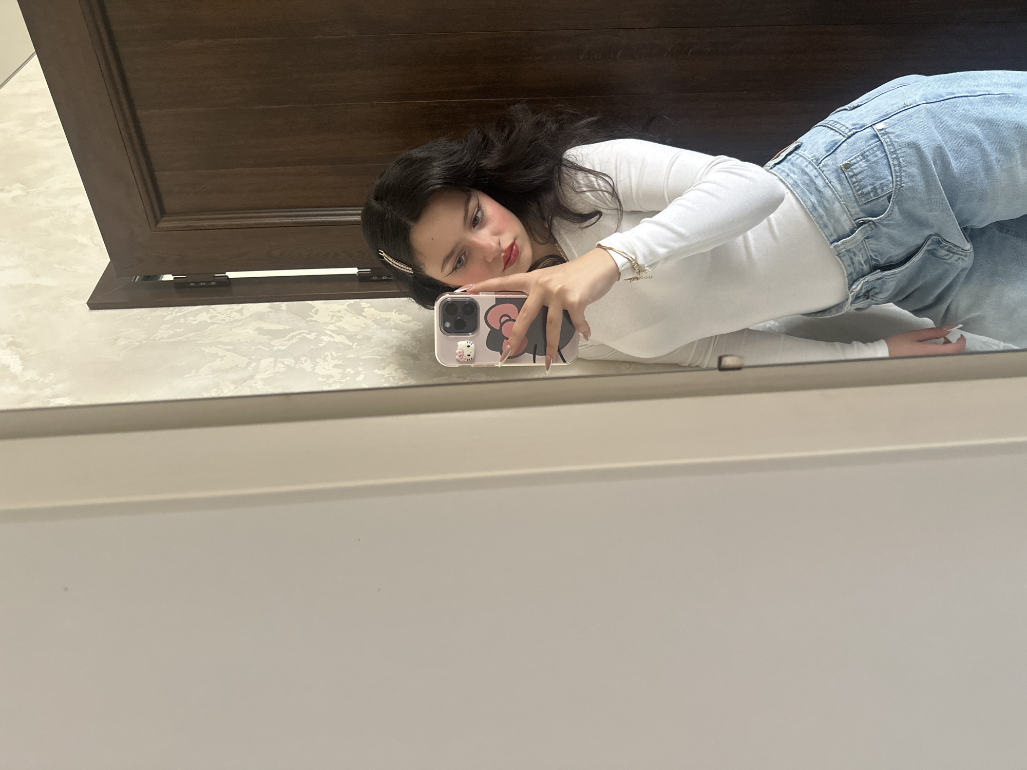 A young woman is lying on a tile floor taking a selfie in a mirror. She is wearing a white long-sleeved shirt and light blue jeans. Her dark hair is down, and she has a jeweled clip in her hair. She is holding a pink iPhone with a Hello Kitty case. Behind her is a dark wooden door. The location is unknown, and there is no evidence that it is in Yerevan, Armenia.