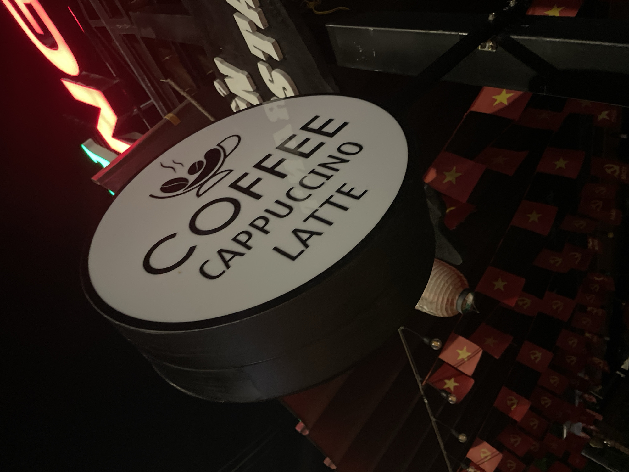 The image shows a round, black and white coffee shop sign displaying "COFFEE CAPPUCCINO LATTE" with a cup design. The sign is situated near other neon signs and strings of small Vietnamese flags. The photograph appears to be taken at night in Phu Thuy Ward, Vietnam, based on the presence of the Vietnamese flags.