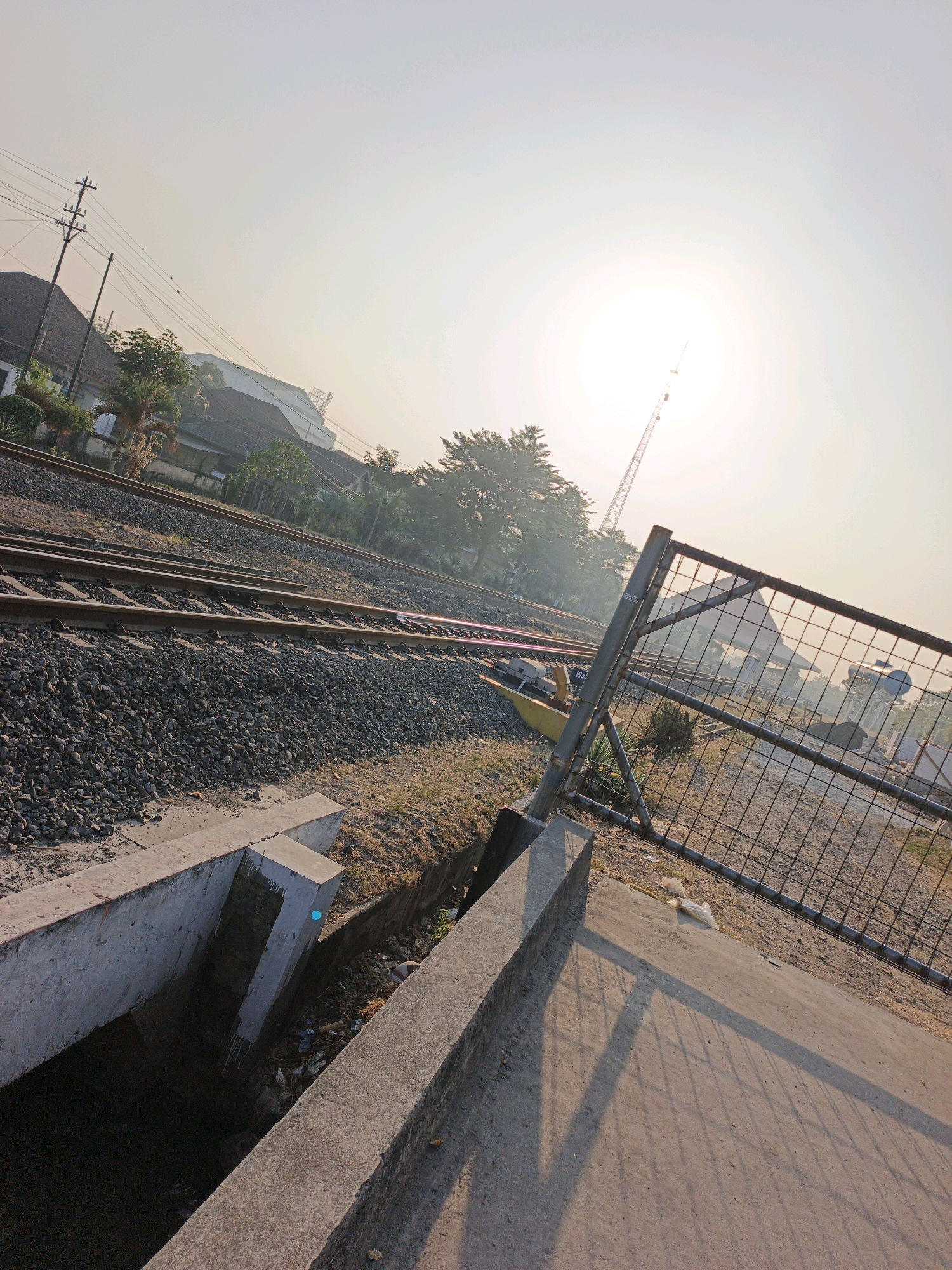 The image depicts a railway track in Mojokerto, Indonesia, likely near a station, under a hazy sky with the sun prominently visible. In the foreground, a concrete drain runs diagonally, filled with some debris. Adjacent to the drain is a concrete ledge and further on, a metal chain-link fence. The railway tracks themselves are laid on a bed of gravel, and they curve into the distance. On the left side of the tracks, buildings and power lines are visible, suggesting an urban or semi-urban environment. A tall, lattice-like structure, possibly a radio tower or construction crane, stands against the bright sky. The overall scene is bathed in the warm light of either sunrise or sunset, with shadows cast by the fence and other objects. There are no people visible in the frame.