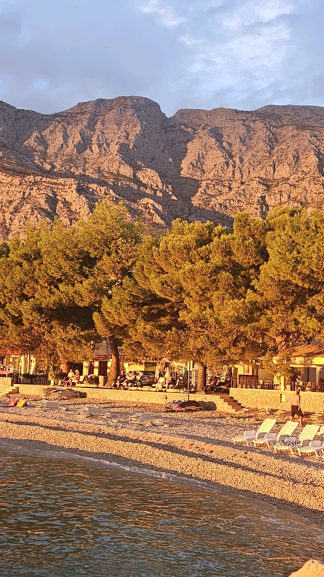 The image depicts a pebble beach in Općina Tučepi, Croatia, with the Biokovo mountain range in the background. The setting sun casts a warm, golden light across the scene. In the foreground, gentle waves lap at the shore. Behind the beach, a row of sun loungers is arranged on the pebbles. Further back, a line of trees with lush green foliage provides shade, with a few people seen sitting or walking amongst them. Beyond the trees, a low building with a stone facade appears to be a restaurant or cafe, with tables and chairs set up outside. Several vehicles, including cars and scooters, are parked along the edge of the boardwalk. The mountains in the background are rugged and rocky, illuminated by the last rays of sunlight, and their textured surfaces are clearly visible. The sky is a soft blue with a few wispy clouds.