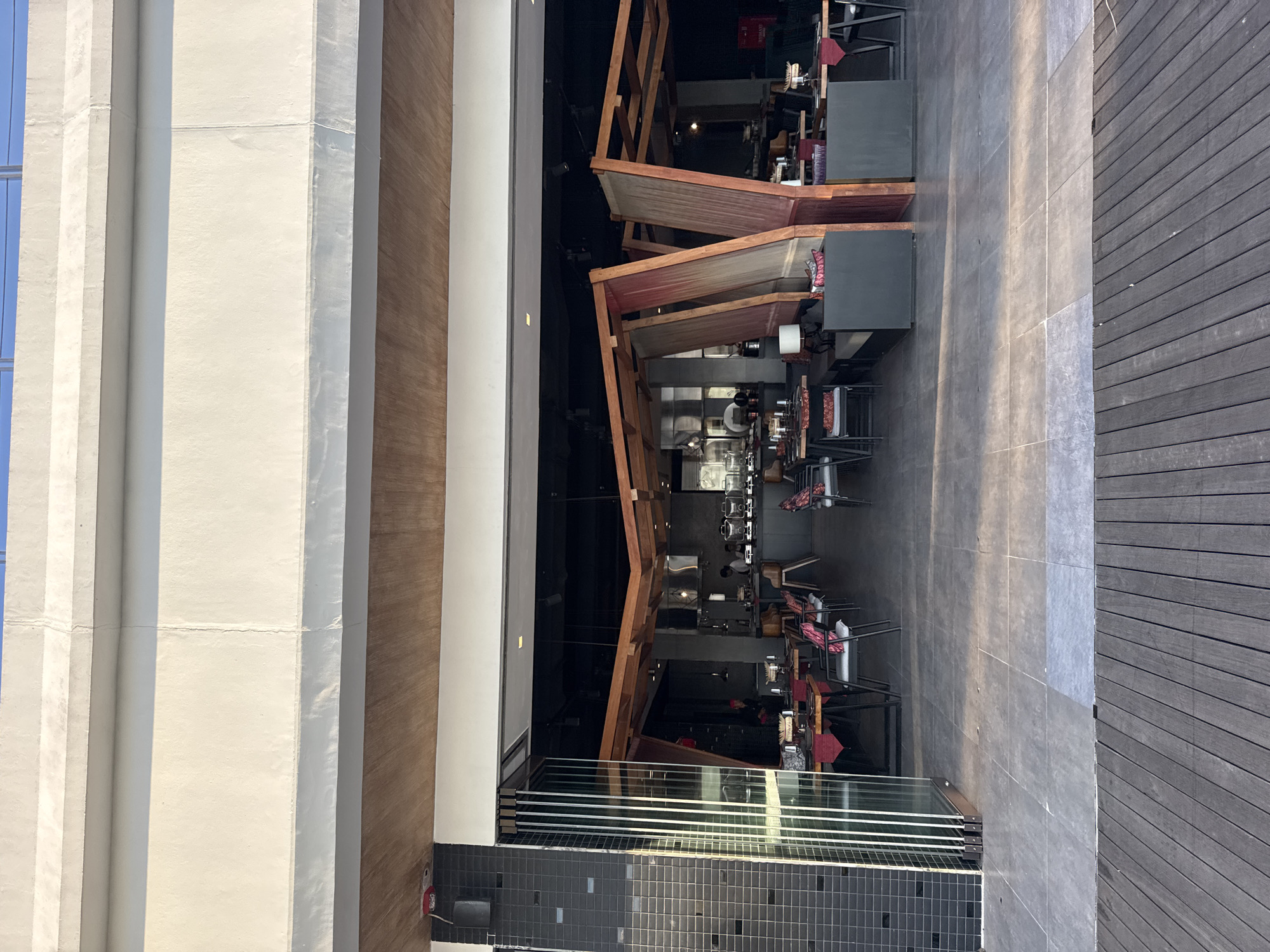 This is an outdoor photograph of a restaurant's entrance in Kuta Selatan, Indonesia, under bright daylight conditions. The restaurant features a modern, open-air design with an interior dining area visible through large glass folding doors. Inside, the restaurant is furnished with tables and chairs, and there is evidence of kitchen activity. The exterior consists of a tiled patio area leading to a wooden deck. The building has a contemporary architectural style, with concrete and wood elements. The sky is visible in the background.