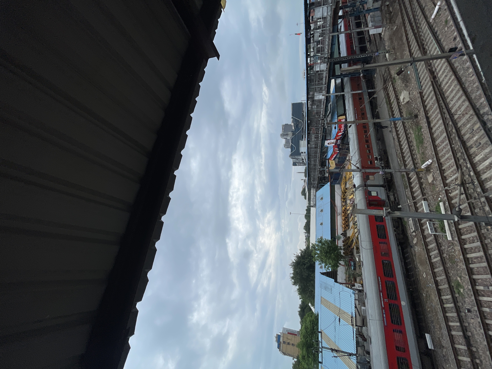 The image captures a view of a train station in New Delhi, India, under a cloudy sky. A corrugated metal roof overhang partially obscures the top of the frame, creating a shadow that emphasizes the contrast with the bright, overcast sky. Below the roof, a train with red and gray carriages is parked on the tracks. The train appears to be at a station platform, with a walkway or bridge visible above it, connecting different parts of the station.
In the background, there are buildings, including a tall, modern structure with multiple levels and windows, and other lower-lying buildings, possibly station facilities or adjacent structures. Trees are scattered throughout the scene, suggesting an urban environment with some greenery. Several sets of train tracks are visible, indicating a busy railway junction. The overall impression is of a functional, perhaps somewhat industrial, urban setting. The image is taken from an elevated perspective, looking down and across the train tracks and platforms.