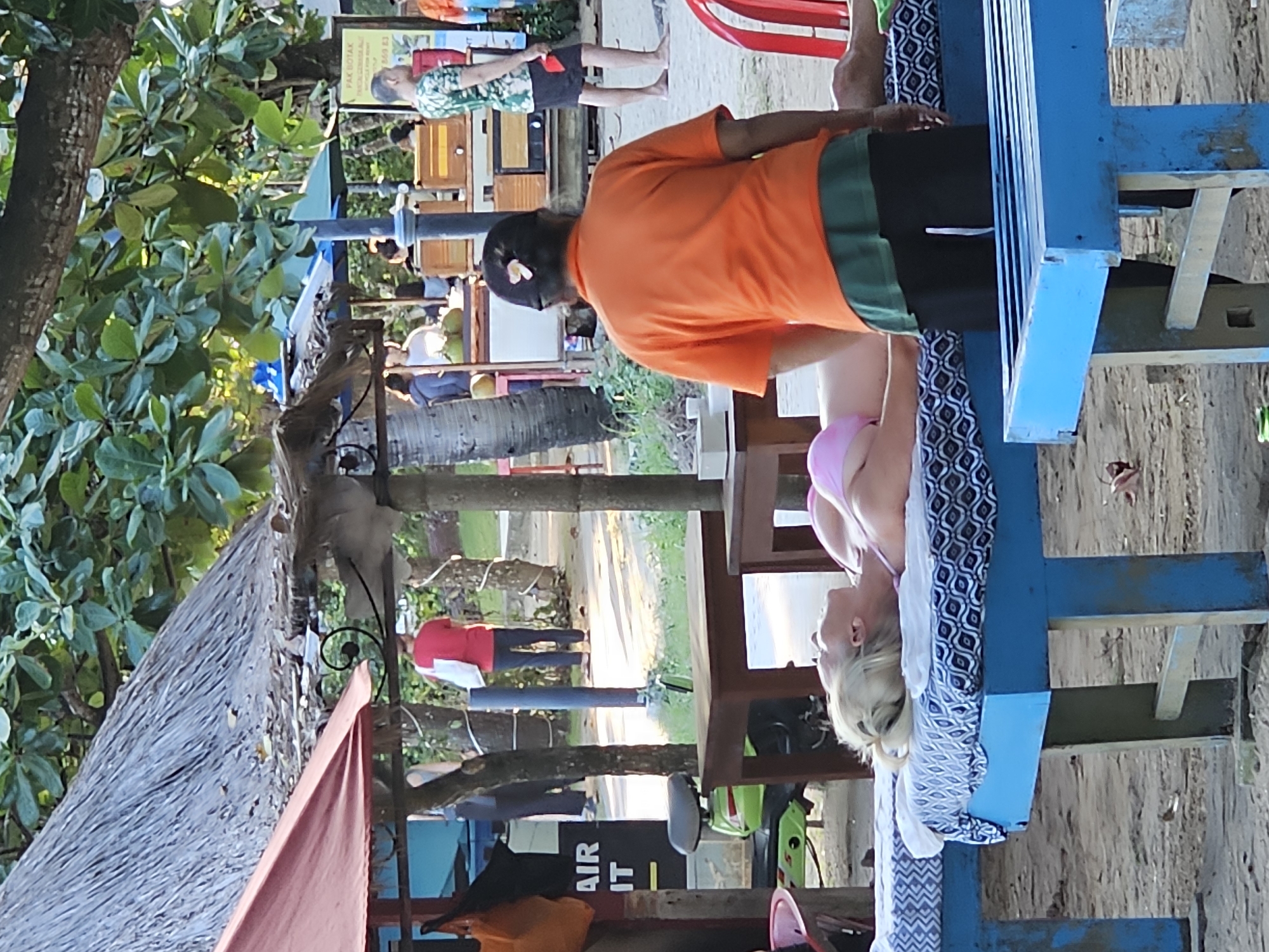 The image captures a scene in Denpasar, Indonesia, featuring a woman in a pink bikini lying on a blue massage table while receiving a massage from a masseur in an orange shirt and black shorts. The massage occurs outdoors, likely in a spa area. A hut with a thatched roof is located nearby. In the background are people, trees, and signage.