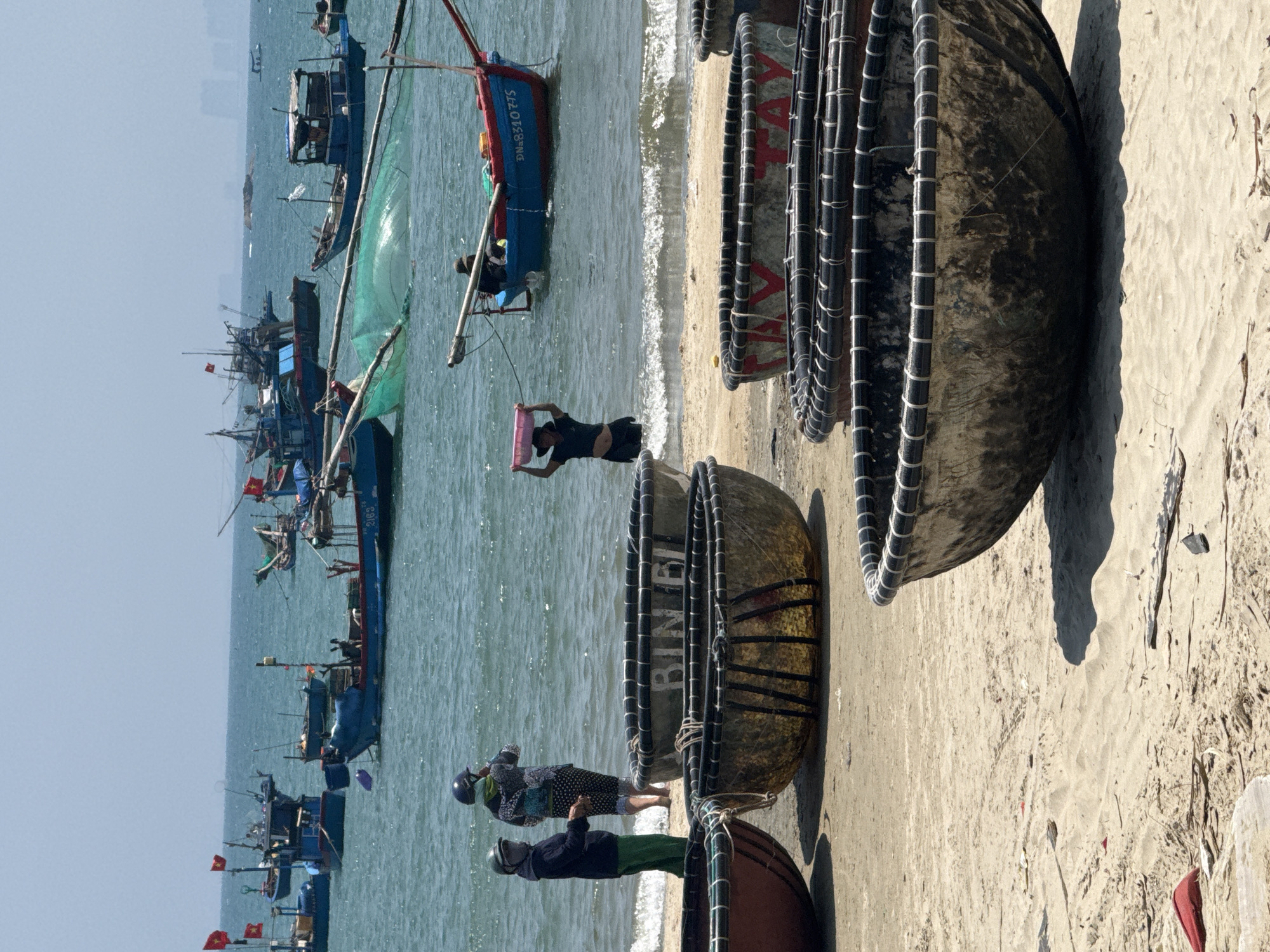 The image shows a beach scene in Đà Nẵng, Vietnam. Several round, woven basket boats are on the sand. The name "BINH DINH" is visible on one boat. People are near the boats and in the water. One person carries a pink container. Fishing boats are visible in the sea. The sky is bright and clear.