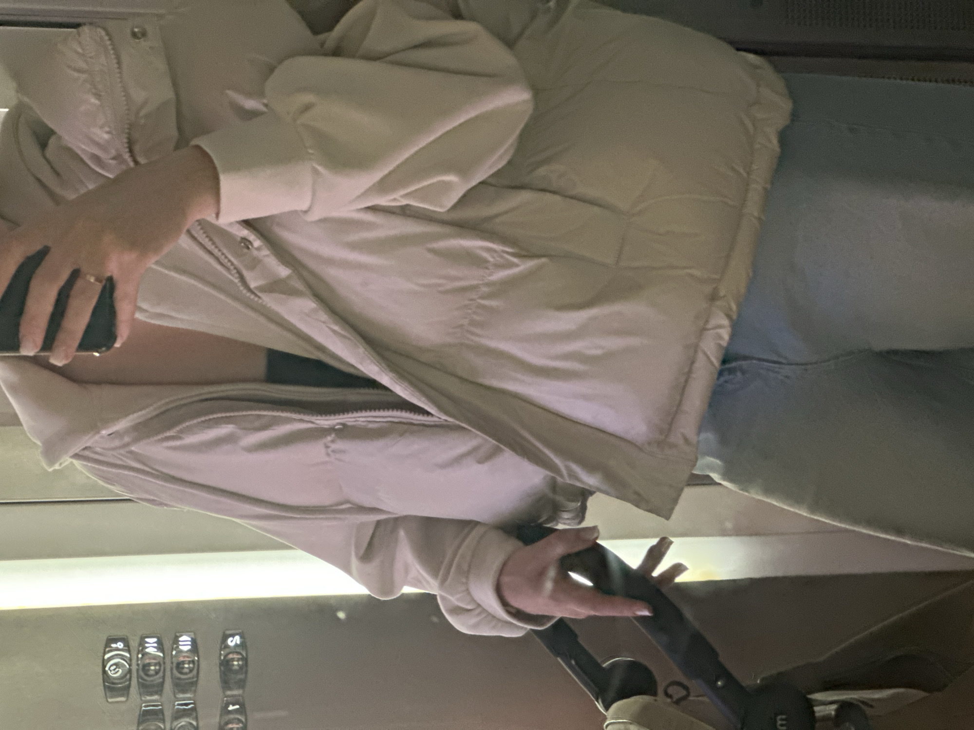 A woman, visible from the mid-torso up, is inside an elevator. She is wearing a beige quilted jacket and light blue jeans. In her left hand, she holds a black smartphone. With her right hand, she is touching or pressing a black elevator control panel. The elevator control panel has raised numeric and alphabetical buttons. The interior of the elevator appears to be stainless steel. The location is assumed to be Kazan, Russia.