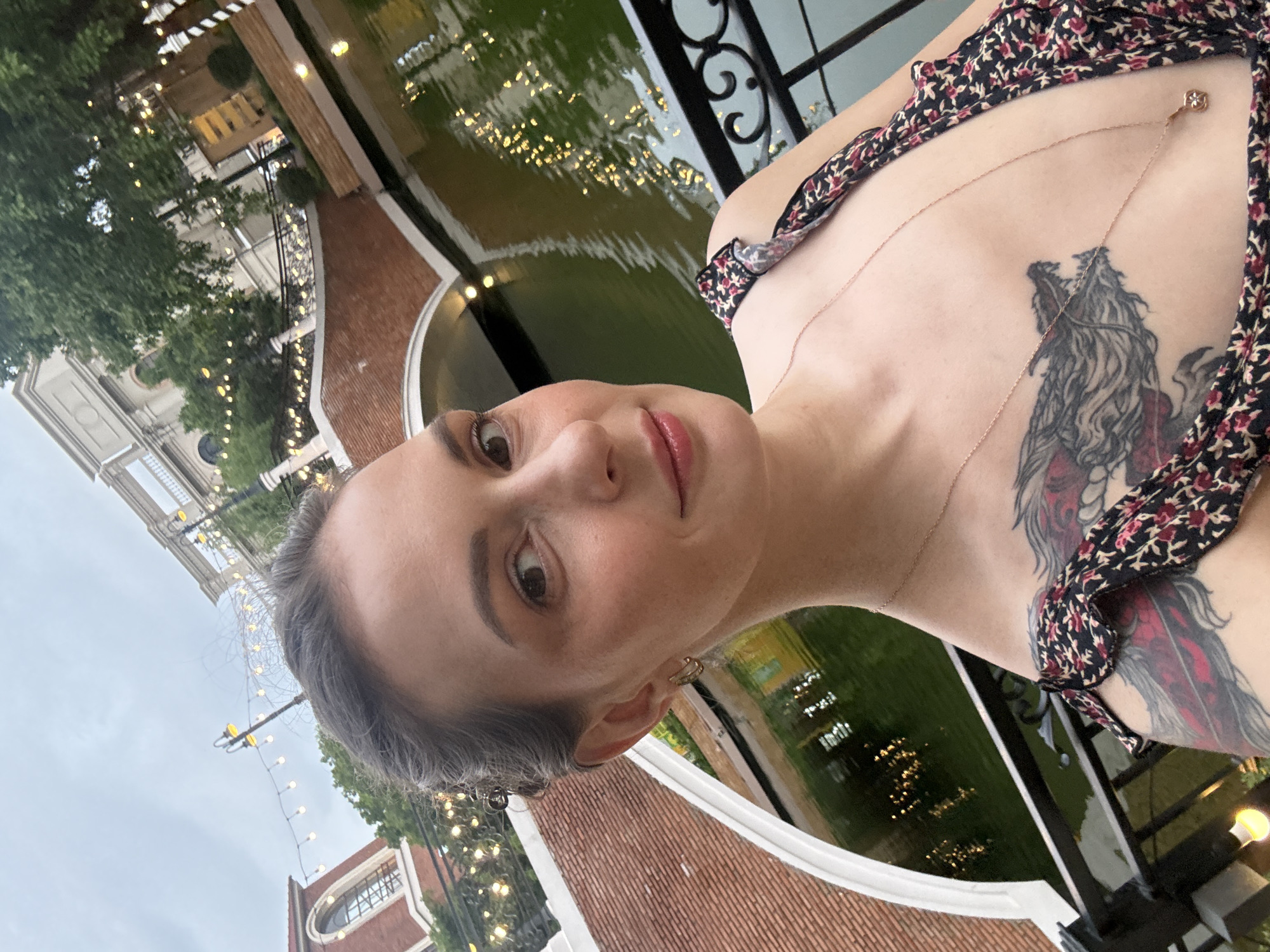 A woman with dark hair pulled back and a dragon tattoo on her chest is taking a selfie. She is wearing a floral print dress and a delicate necklace. The background shows a canal with reflections of lights, a brick bridge, and buildings with ornate architecture, suggesting a European-inspired setting. The presence of string lights indicates it is likely evening or nighttime. The location is identified as the city of Khan Chbar Ampov, Cambodia.