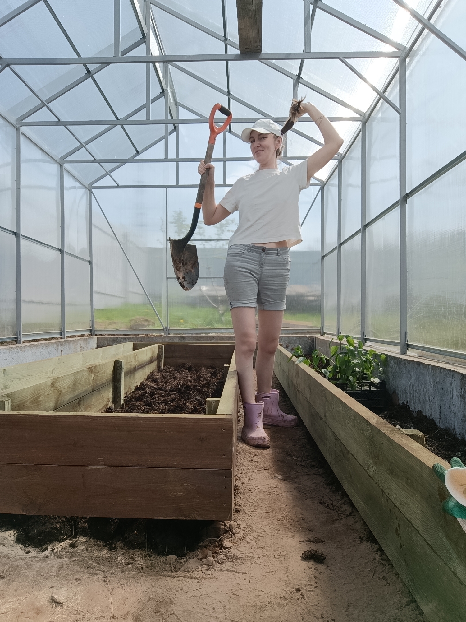 The photograph depicts a young woman standing inside a greenhouse in Chaikovsky, Russia. She is holding a shovel over her shoulder and playfully pulling at her hair. She wears a white t-shirt, grey shorts, a white baseball cap, and pink rubber boots. The greenhouse has a metal frame with translucent panels. Inside, there are two raised garden beds. One bed is filled with dark soil, while the other appears to contain small plants. The ground is dirt. Sunlight shines through the roof of the greenhouse.
