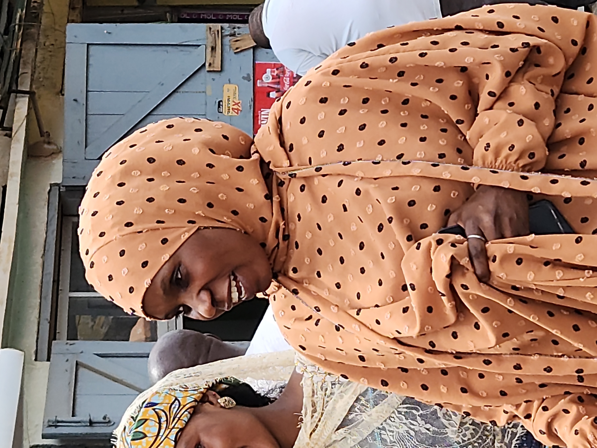 In a medium shot, a woman wearing a peach-colored headscarf with black polka dots and a matching patterned dress smiles. She has dark skin and her hair is not visible. Her dress has a gathered neckline and billowing sleeves. To her left, another woman, partially visible, wears a yellow and white patterned headscarf and a cream-colored lace dress. Behind them, a blue wooden door with a diagonal plank and a metal handle is set into a weathered building. A partially obscured sign with "4X" is affixed to the door. A man in a white shirt is visible to the right, his back to the camera, and another person with a bald head stands between the two women. The environment appears to be outdoors in a bustling urban setting.