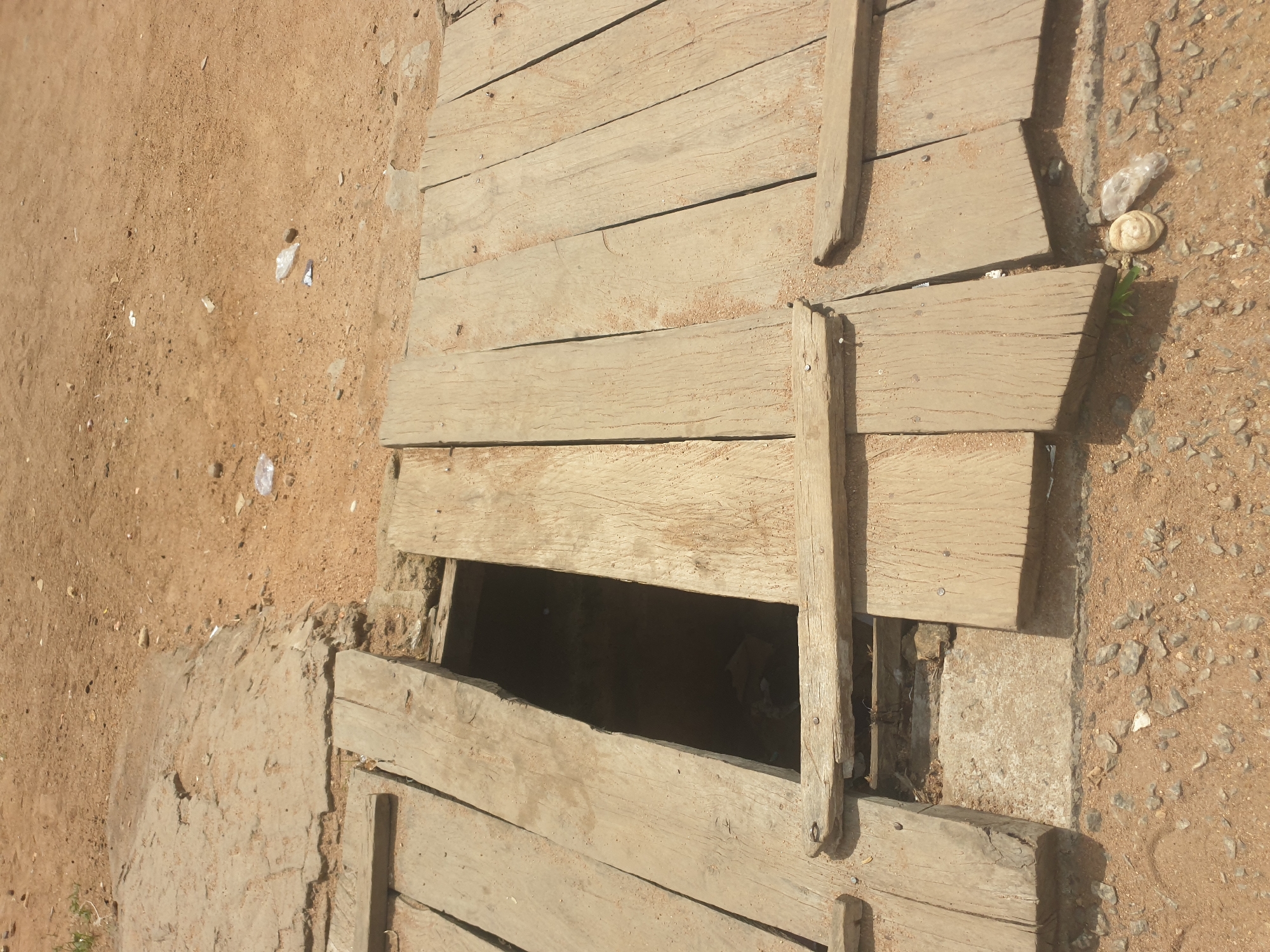 A close-up, overhead view shows a dilapidated wooden walkway over a dark, recessed area. The walkway is constructed of weathered, splintered wooden planks, some of which are unevenly spaced or broken, revealing the darkness beneath. The surrounding ground is a mix of dry, sandy soil, gravel, and some scattered debris, including a discarded plastic bottle and a small, round object. The overall impression is one of disrepair and neglect.