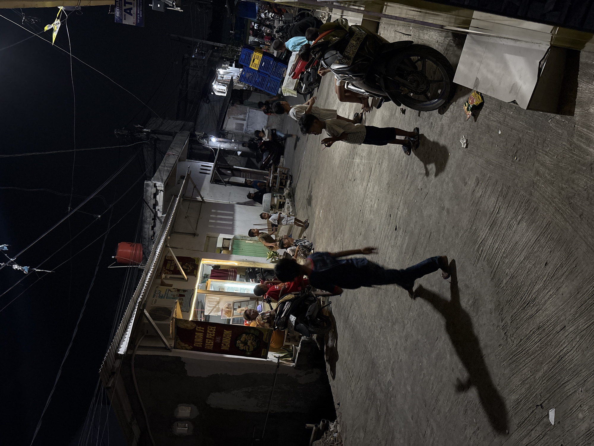 In North Jakarta, Indonesia, at night, a group of children are gathered on a street in front of a brightly lit food stall. The stall, marked with Indonesian text indicating it sells "Nasi Pecel" (rice with mixed vegetables and peanut sauce), has a glass display case showing food items. Several motorcycles are parked on the street, some in front of the stall and others further down the road. A few adults are also present, including one sitting on a motorcycle near the stall and another standing in the background. The street is paved with concrete and has overhead electrical wires and poles. There are various structures lining the street, including buildings and what appears to be an ATM booth visible in the distance. The scene suggests a casual, community gathering occurring in the evening.