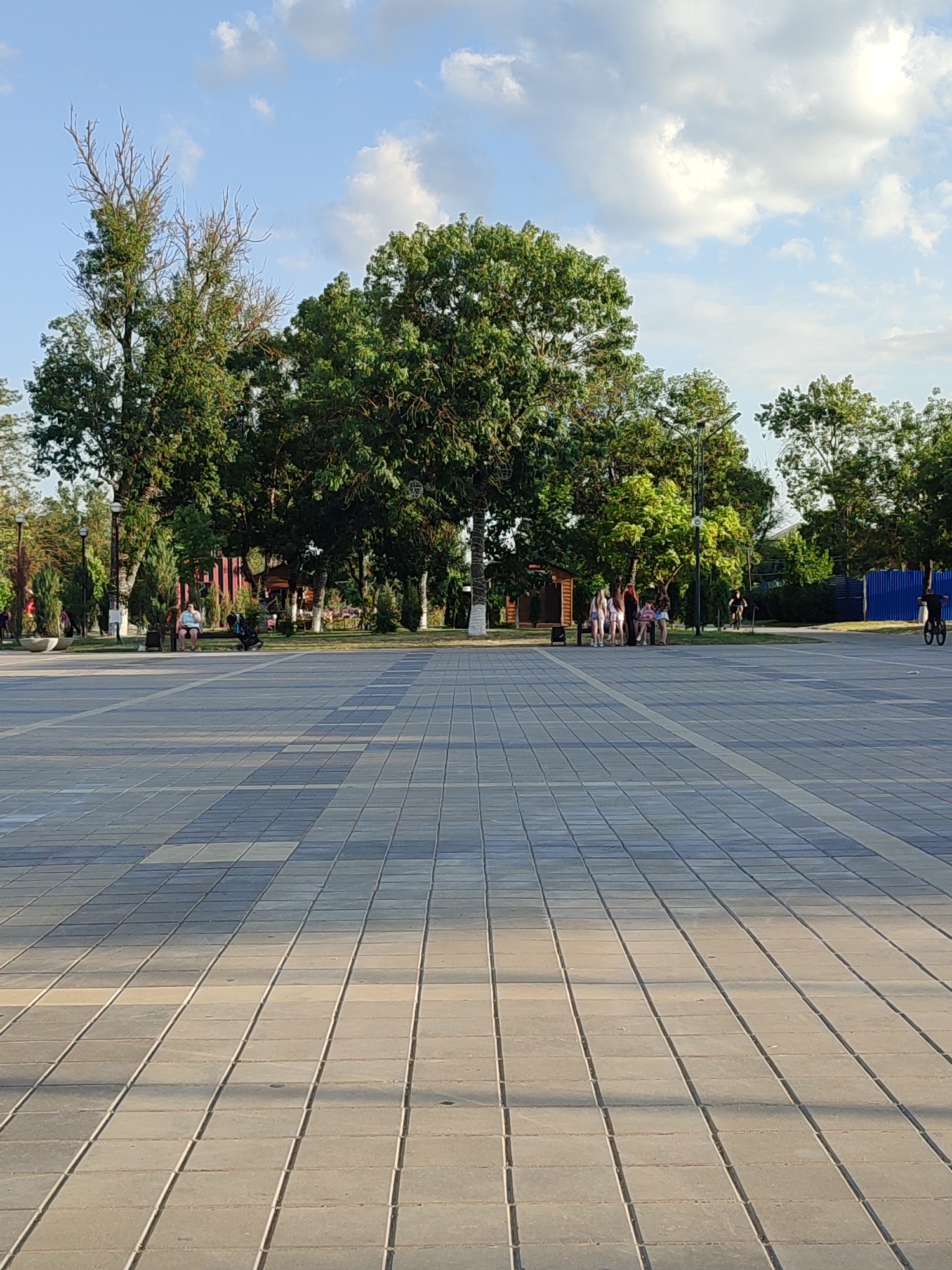 The image depicts a wide, paved plaza in a city park. The plaza is laid with rectangular grey tiles arranged in a grid pattern, with some tiles reflecting sunlight, creating alternating light and dark bands. In the background, a line of trees with lush green foliage stands along the edge of the plaza, with a few older trees showing bare branches. Beyond the trees, there are structures that appear to be part of a recreational area, including wooden buildings with red roofs and picnic tables. Several people are visible in the middle ground. On the left, a person sits on a bench next to a stroller. In the center, a group of young people, possibly teenagers, are gathered. Further to the right, another person rides a bicycle. The sky above is blue with scattered white clouds. The overall scene suggests a pleasant day in a public park located in Abinsk, Russia.