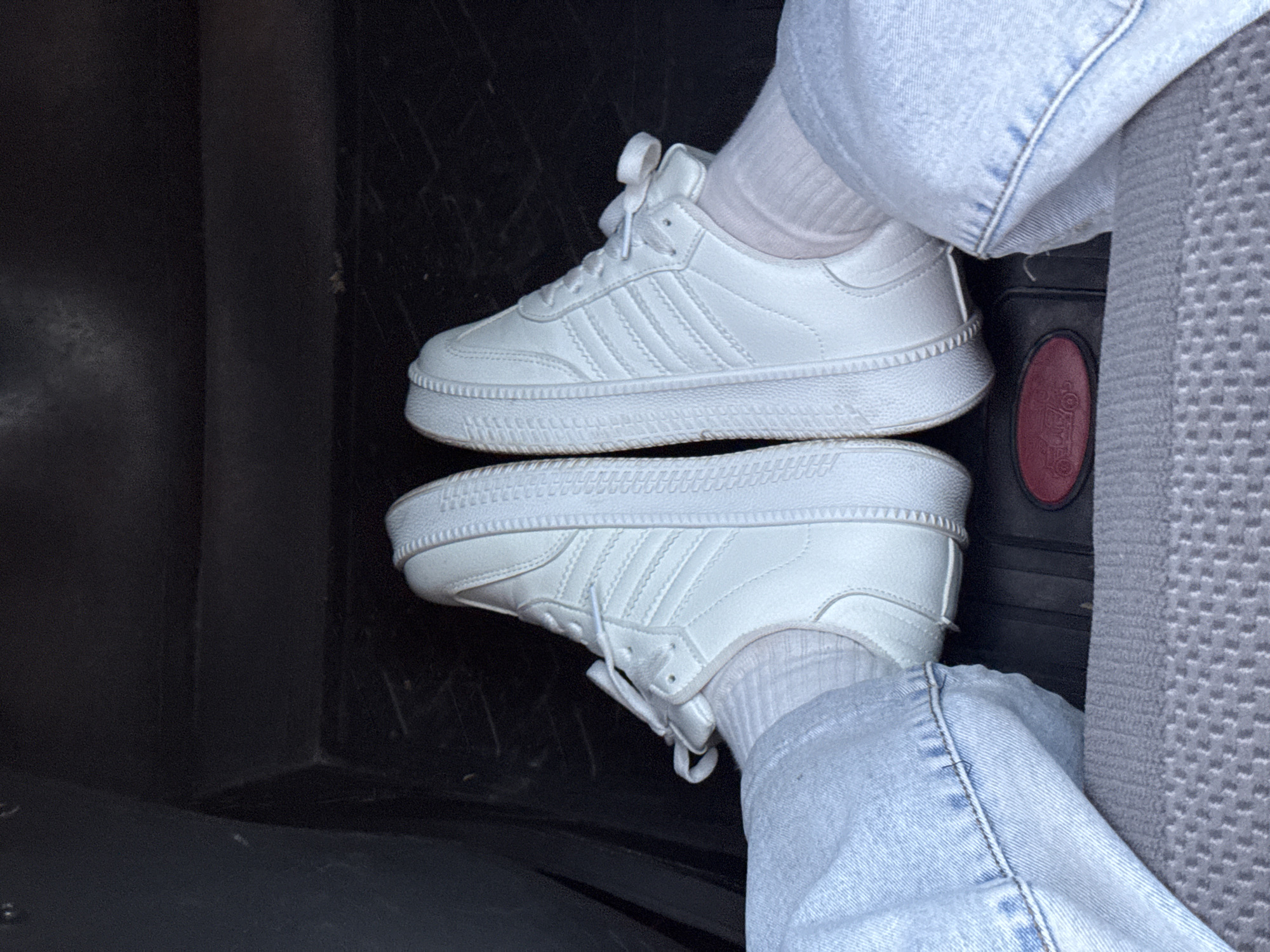 The image shows a pair of white sneakers stacked on top of each other inside a dark compartment, likely within a vehicle. The sneakers appear to be a similar design, with the top one slightly angled towards the viewer. The wearer is wearing light blue denim jeans and white socks. A portion of a gray patterned car seat is visible on the right side of the image. The black compartment appears to be in the floor. The location of Bizyar, Russia, is not evident in the image.