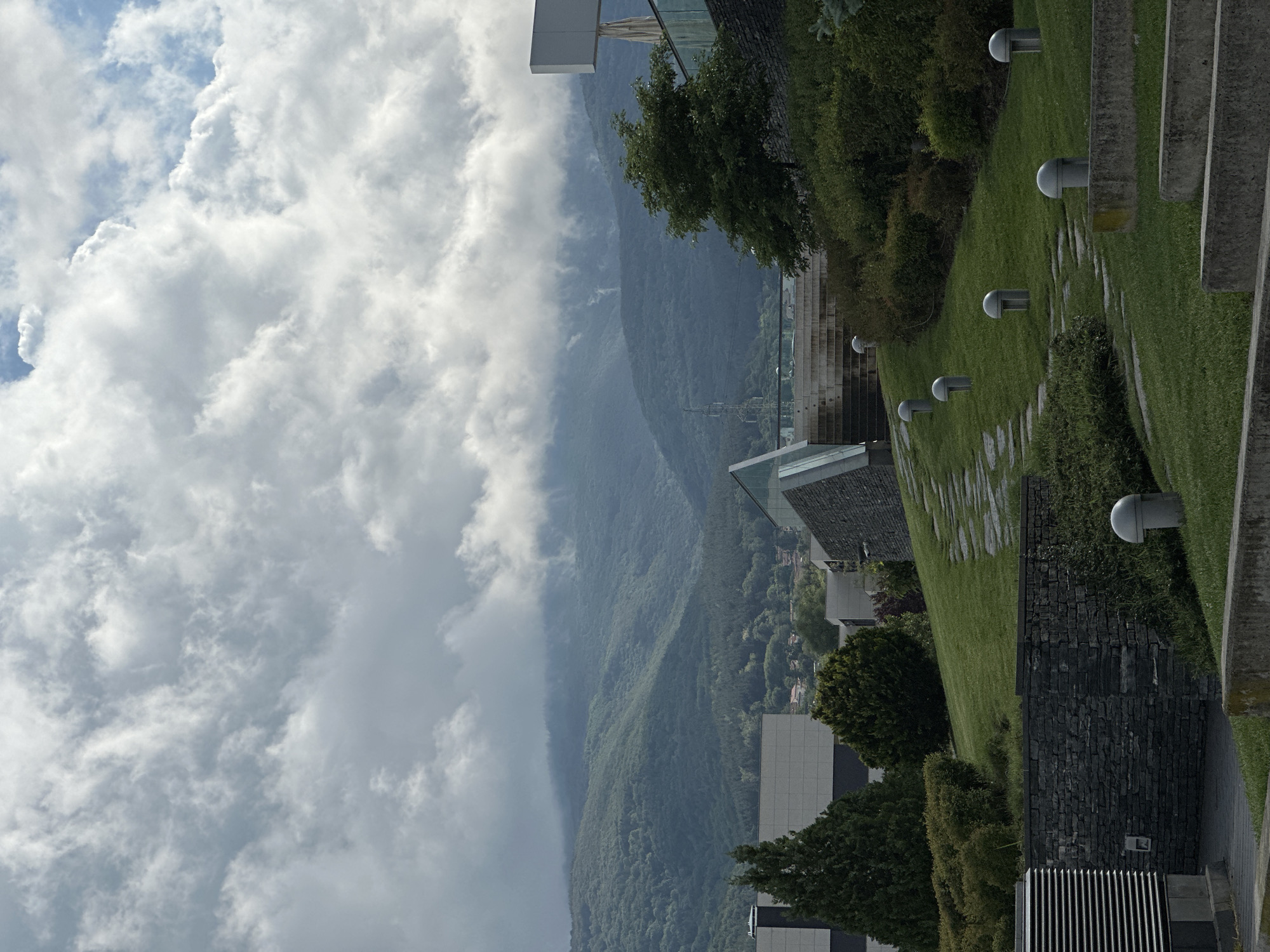 The image captures a scenic panoramic view combining modern architecture with a majestic mountainous landscape, likely situated near Sofia, Bulgaria.
In the upper section of the frame, a dynamic sky is filled with voluminous white and grey cumulus clouds, suggesting a partly cloudy or atmospheric day. Below this, a vast expanse of lush, green mountains stretches across the midground. These mountains are densely covered in forests, and parts of their peaks and slopes are softened by a layer of haze or low-hanging clouds, adding depth to the scene. Faint glimpses of a distant town or city can be seen nestled at the base of the mountains.
The foreground features contemporary architectural elements and carefully landscaped grounds. A vibrant green lawn slopes gently downwards from the right, incorporating irregular stone pathways. Several modern, spherical-topped light fixtures are embedded along these paths and the grassy areas. Dark green bushes and trees are strategically placed, enhancing the modern aesthetic of the built environment. Structures with sleek glass elements and dark stone or concrete cladding are visible, suggesting modern buildings, possibly part of a resort, park, or upscale development. A rectangular sign or structure can be observed on top of one building on the far left.
The setting appears to be daytime, likely late morning or early afternoon, characterized by bright but diffused light due to the cloud cover. The overall impression is one of tranquility and thoughtful design integrated with nature. There are no people visible, and no discernible text is present in the image.