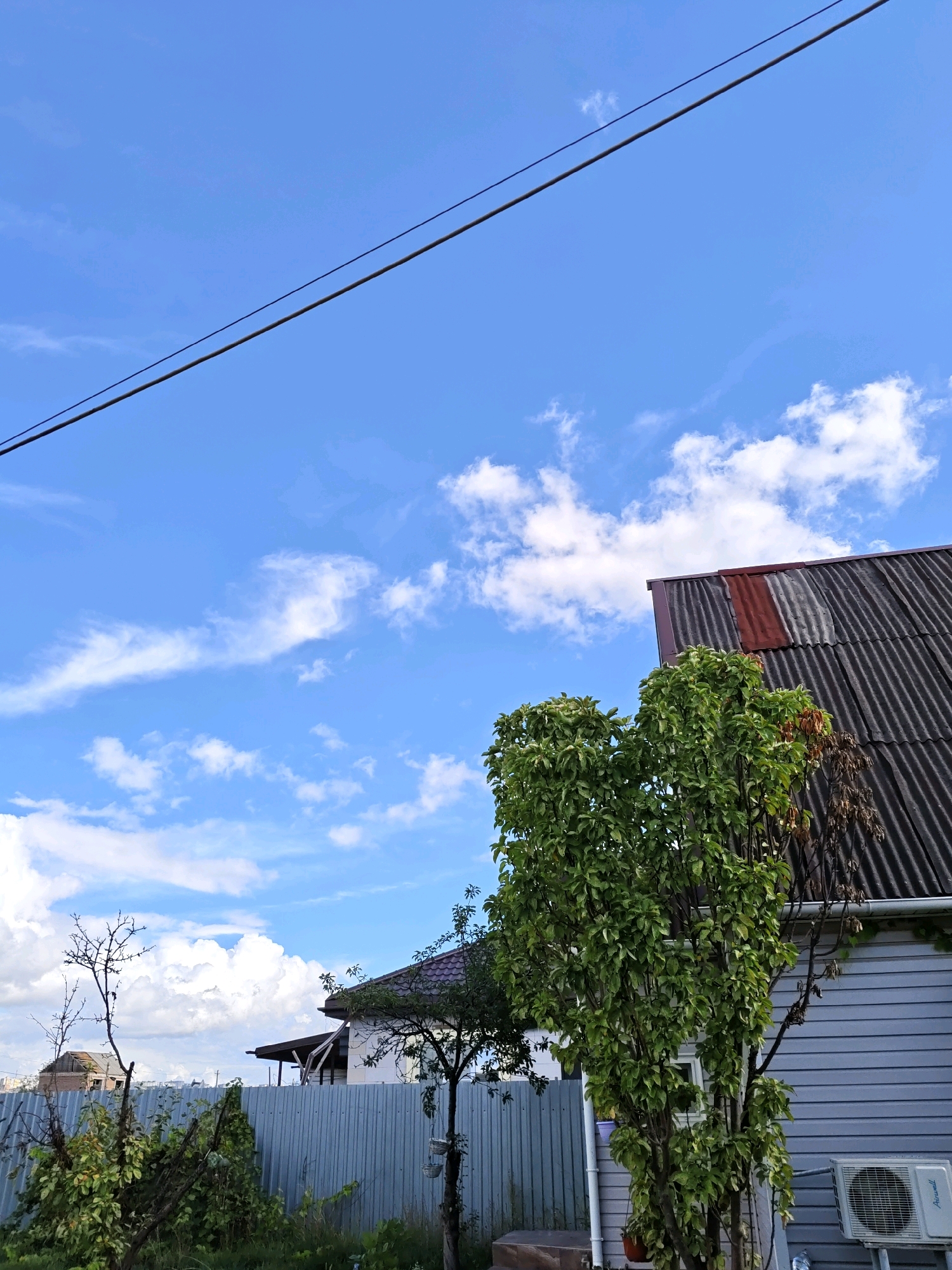 The image depicts a residential area with houses and a fence under a bright blue sky with scattered white clouds. Two parallel black electrical wires are visible stretching diagonally across the upper portion of the frame. In the foreground, a tall, leafy green tree is prominently featured. To the right of the tree, a gray house with a corrugated, dark-colored roof is visible, with a red patch on the roof. An outdoor air conditioning unit is mounted on the side of the house. To the left, a lighter-colored house with a dark tiled roof is partially visible behind a gray metal fence. A bare-branched tree stands to the left of the fence. The scene suggests a daytime view in a suburban or rural setting. The location is identified as Rostov-on-Don, Russia.