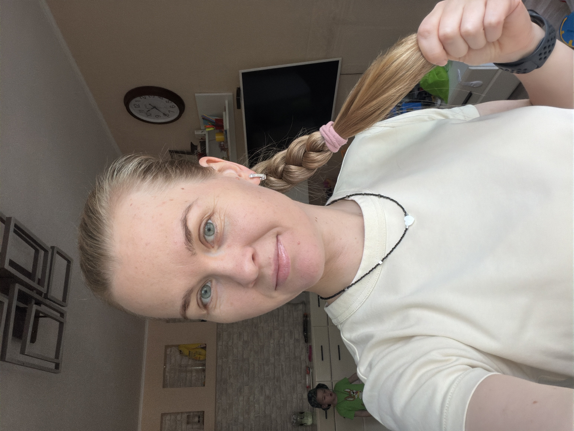 The image is a selfie of a woman with blonde hair in a braid, wearing a light-colored t-shirt and a black beaded necklace. She has blue eyes and is smiling slightly. In the background, there is a clock on the wall, a television, a shelf with items, and a child in a green shirt standing near a dresser. The room appears to be a living space or bedroom. The image metadata indicates it was taken in Khabarovsk, Russia.