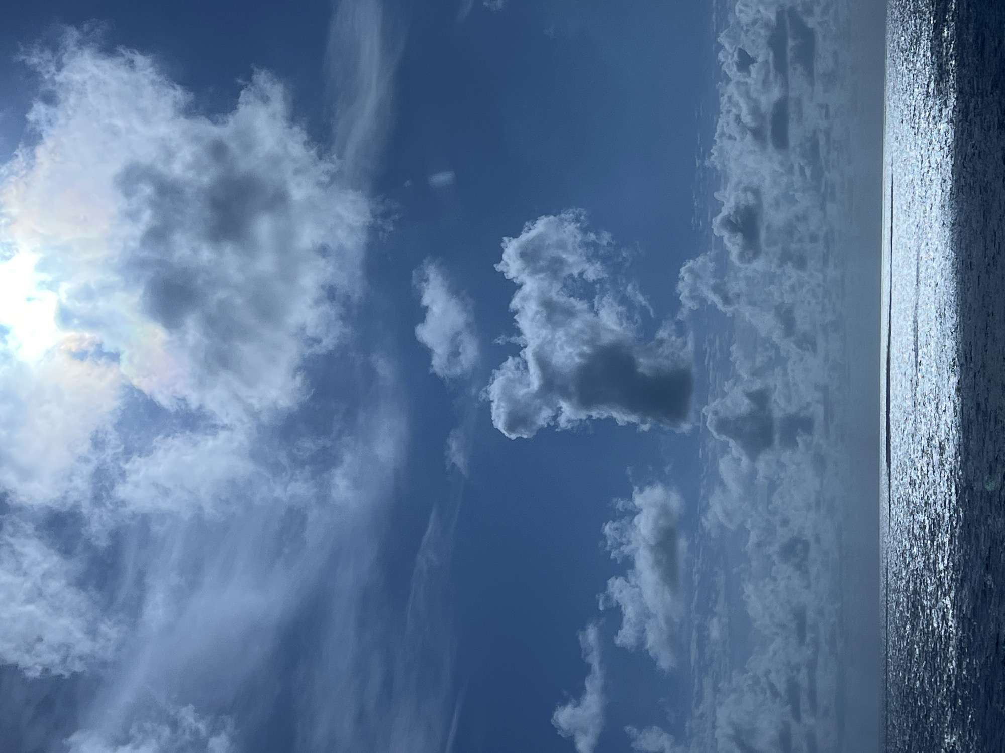 A photograph captures a vast expanse of a deep blue sky filled with dramatic, textured clouds, ranging from wispy cirrus to darker, more dense formations. The sun is partially obscured by a large, bright cloud formation in the upper right quadrant of the image, creating a luminous glow and a subtle rainbow effect. Below the sky, a dark, tranquil sea stretches to the horizon, its surface shimmering with reflected sunlight, creating a bright, horizontal band of light. The overall impression is one of a dynamic and serene seascape, likely taken from a boat or a coastal viewpoint. The location is identified as Unknown, Unknown.