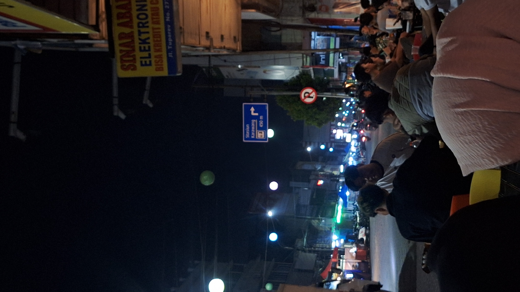 The image shows a street scene at night in Karawang, Indonesia. A sign indicates that the Karawang train station is 450 meters away. Another sign advertises "Sinar Abadi Electronics" at Jl. Tuparev No. 277. People are seated, possibly at a restaurant, and several motorcycles are visible in the background. The illumination comes from streetlights and the lights of various businesses. A no-right-turn sign is also present.