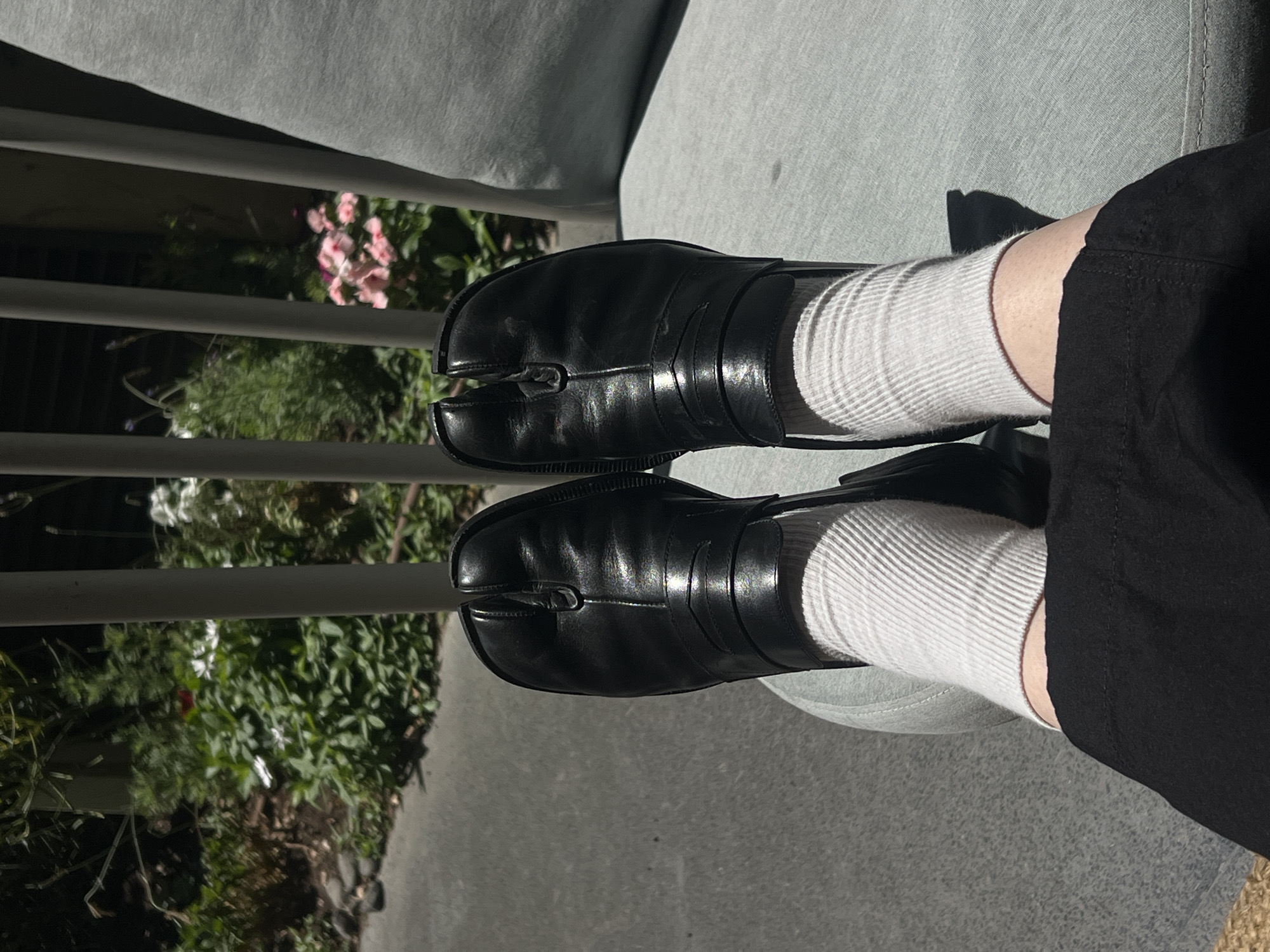 A person's legs are crossed and angled toward the camera, resting on an outdoor chair. They are wearing black tabi-style shoes and white ribbed socks. The background shows a lush green garden with some pink and white flowers, and a white railing. The flooring is a textured gray surface, possibly a patio or walkway. The overall impression is one of relaxed outdoor leisure, possibly in a garden setting within Tel Aviv, Israel.