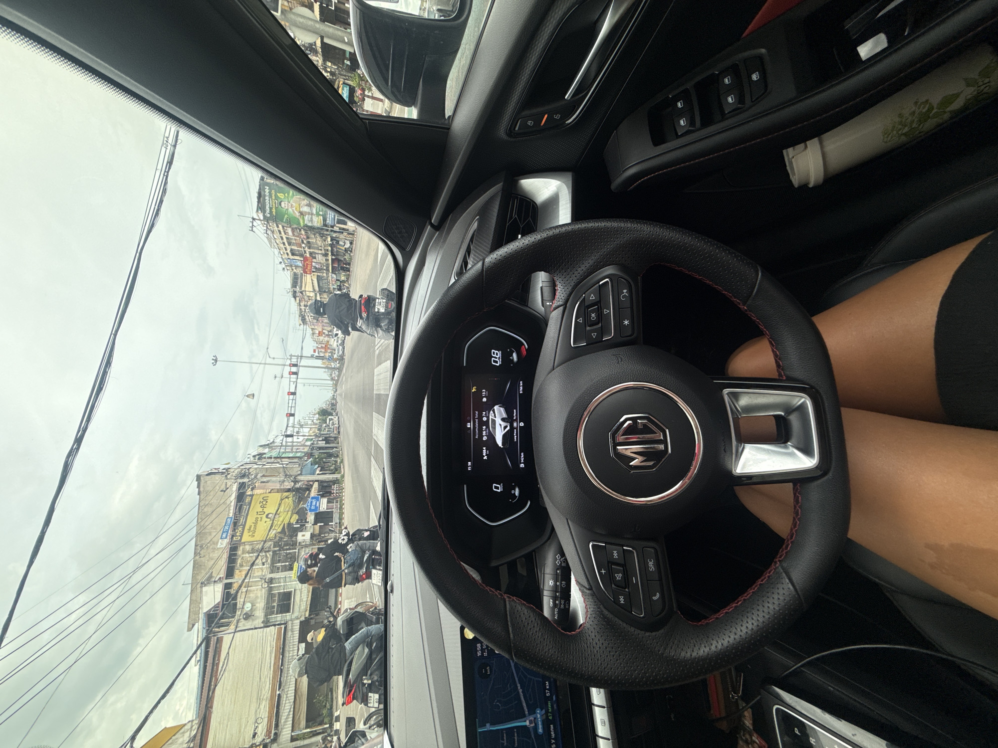 The image is a first-person perspective from inside a car, looking forward through the windshield. The car appears to be stopped at a traffic light on a street in Pran Buri, Thailand. The street is lined with two- and three-story buildings, some with awnings and signage in Thai script. Several motorbikes are visible on the road, with riders wearing helmets. Overhead, a complex network of electrical wires crisscrosses the sky, which is overcast. The car's dashboard displays are active, showing navigation information on the left screen and vehicle data on the right. The steering wheel, with the MG logo in the center, is prominent in the foreground, and a person's legs are visible, indicating they are the driver. A water bottle is also seen in the car's door pocket. The overall scene depicts a typical urban streetscape in Thailand under a cloudy sky.