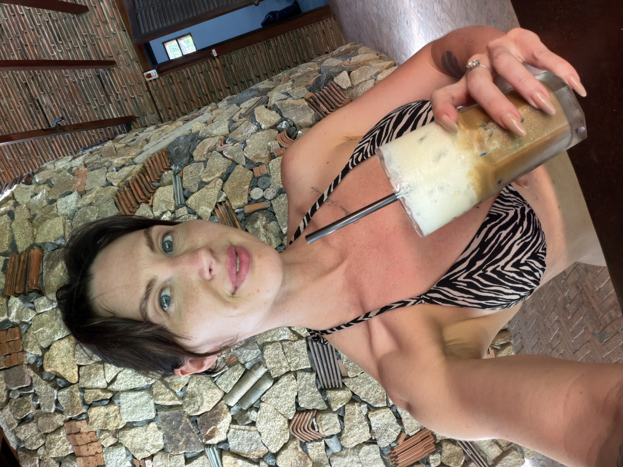 A woman with medium-brown hair pulled back and light skin is holding a tall glass of iced coffee with a black straw. She is wearing a black and white zebra-print bikini top. Her left arm is extended towards the camera, and her right hand is holding the glass. She is smiling and looking at the camera. The background consists of a rough stone wall and a wooden roof structure. Part of a doorway is visible in the upper right corner. The location appears to be outdoors, possibly a cafe or resort in Tay Nha Trang Ward, Vietnam.