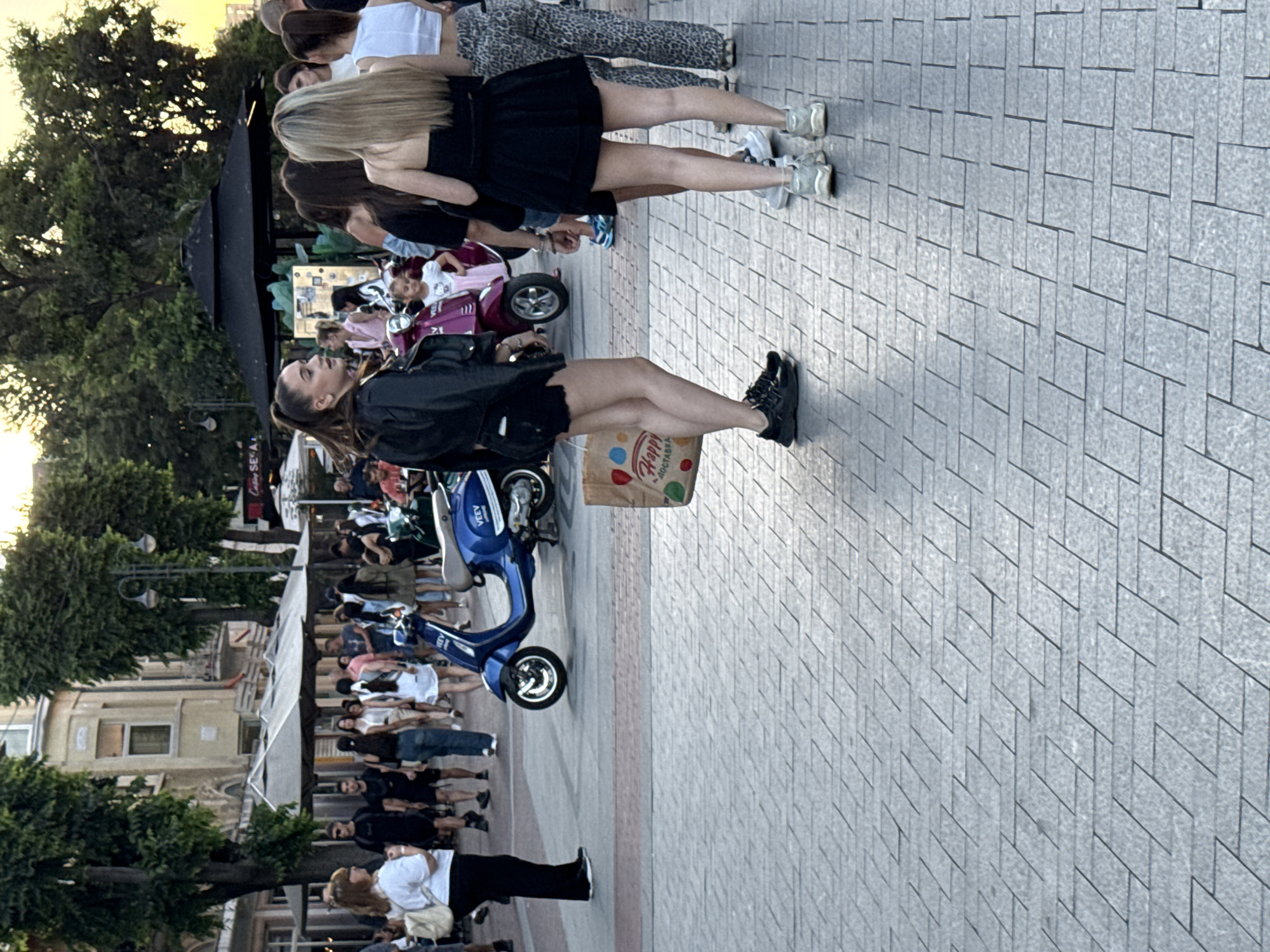 A street scene in an urban environment shows a group of young adults, mostly women, gathered on a paved sidewalk. The people are casually dressed, with some wearing shorts and t-shirts, while others are in dresses or skirts. A blue scooter is parked in the background, and there are trees and buildings lining the street. A paper bag with a "Happy" logo and some fruit illustrations is being carried by one of the women. The lighting suggests it is late afternoon or early evening.