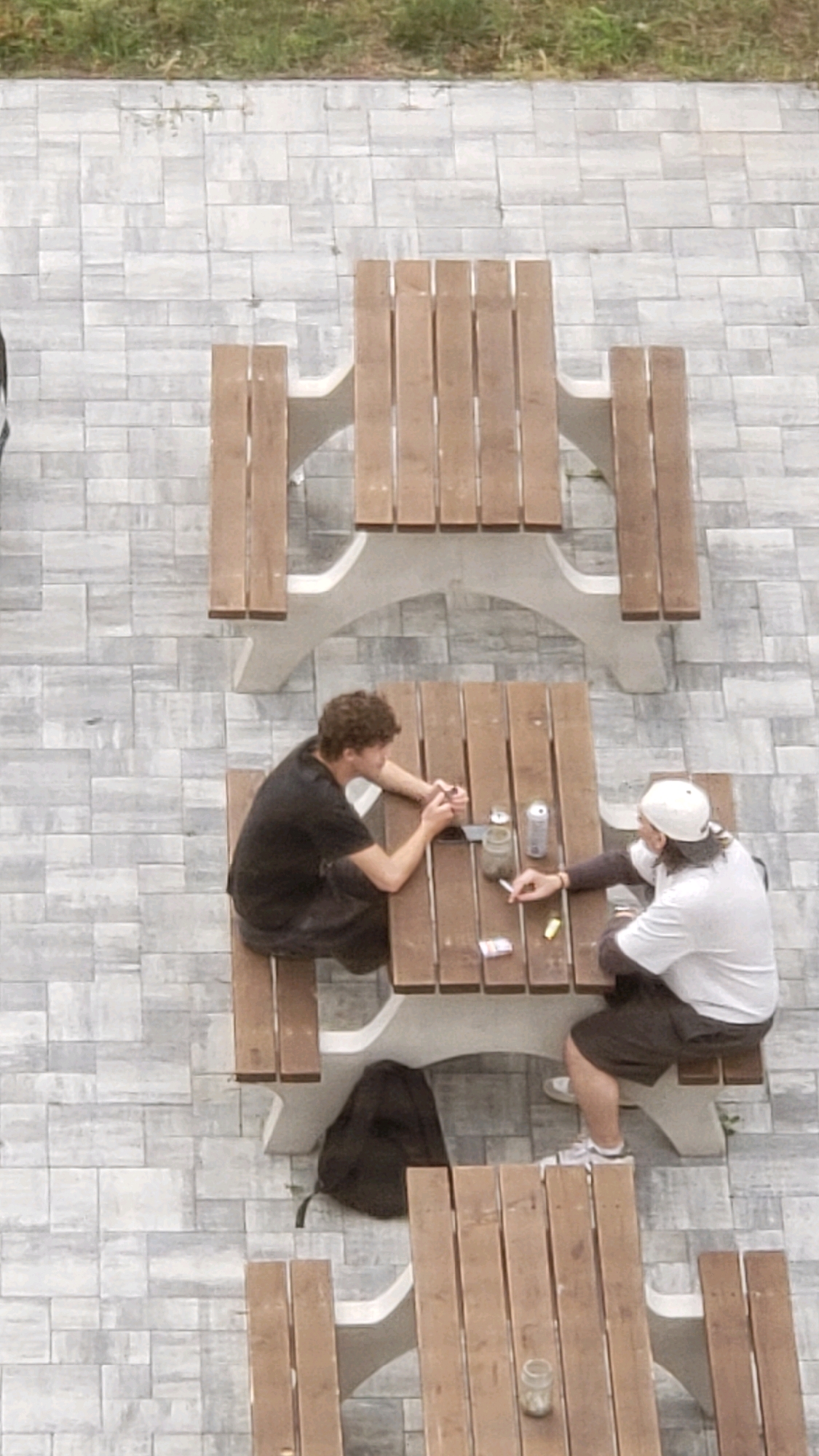 Two individuals are seated at a wooden picnic table on a paved outdoor patio in Prešov, Slovakia. The individual on the left, wearing a black t-shirt and dark shorts, is facing the table with their elbows on the surface and hands clasped. The individual on the right, wearing a white t-shirt, dark shorts, and a white baseball cap, is also facing the table. There are several objects on the table, including two cans, a jar, a cigarette, and a pack of what appears to be cigarettes. A dark backpack is placed on the ground near the picnic table. The surrounding environment consists of a grey, patterned paved surface and a strip of green grass at the top of the frame. The perspective is from above, looking down at the scene.