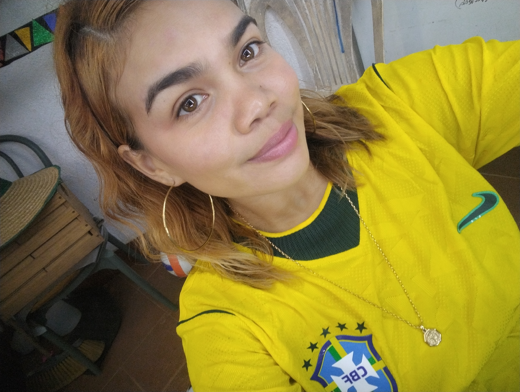 A young woman with blonde-brown hair is looking at the camera in this close-up, high-angle shot taken indoors. She is wearing a yellow jersey with green trim, a gold chain, and large hoop earrings. The background shows a wall with colorful triangular decorations and a portion of a wooden chair with a woven seat. The overall impression is a casual, selfie-style portrait.