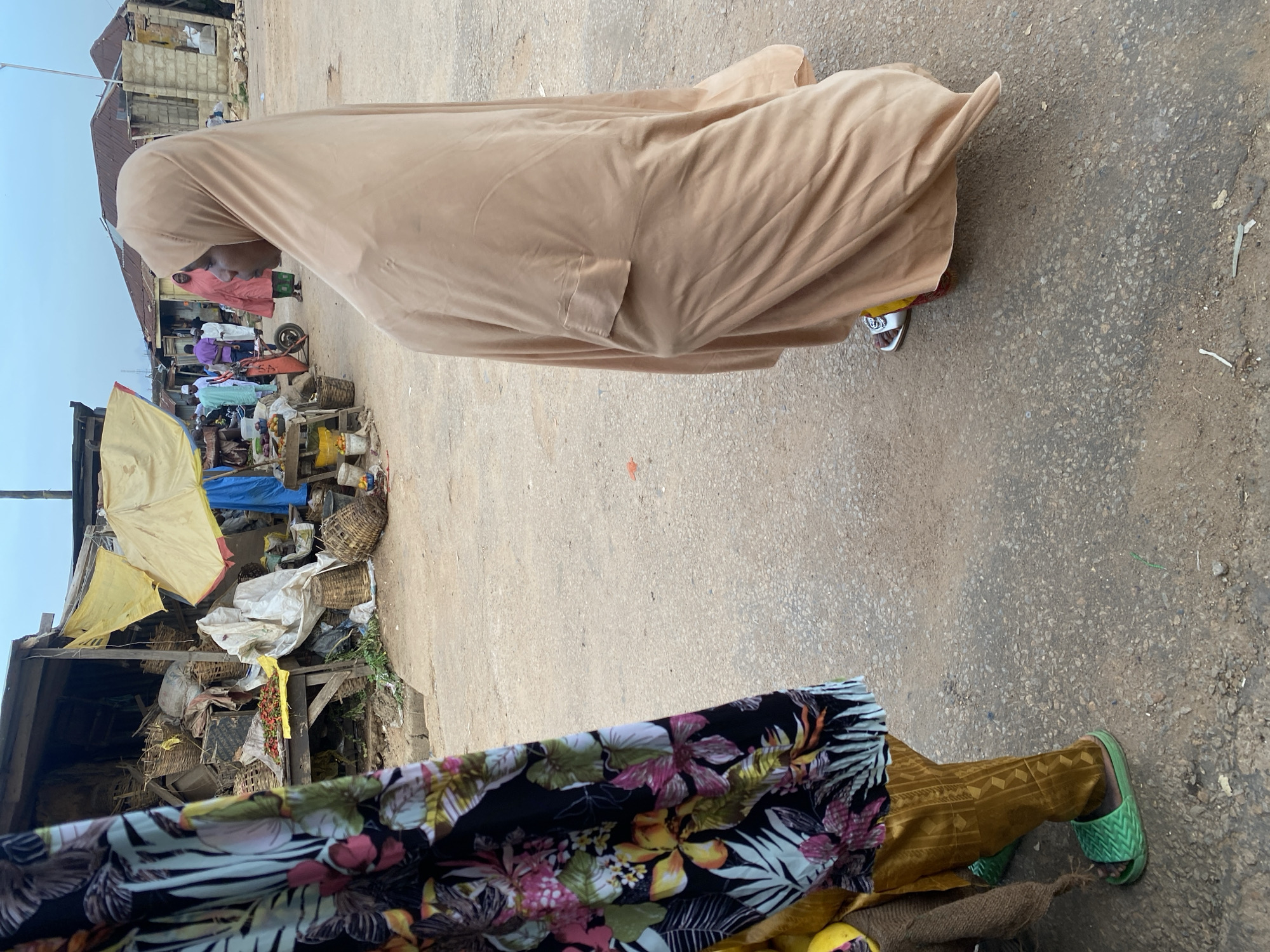 In Jos, Nigeria, a busy marketplace scene unfolds. In the foreground, a person's legs are visible, clad in golden patterned trousers and bright green slide sandals. They are partially obscured by a vibrant floral-patterned garment. To the right, a woman draped in a flowing, light brown hijab stands facing away from the camera, her gaze directed towards the market stalls. Her attire suggests a modest style of dress. The marketplace is filled with various stalls, some covered with yellow awnings, displaying a variety of goods, including baskets and produce. The ground is paved with asphalt, showing signs of wear and tear. In the background, other individuals are seen moving through the market, some in traditional clothing. The overall atmosphere appears to be one of active commerce and daily life in a Nigerian city.