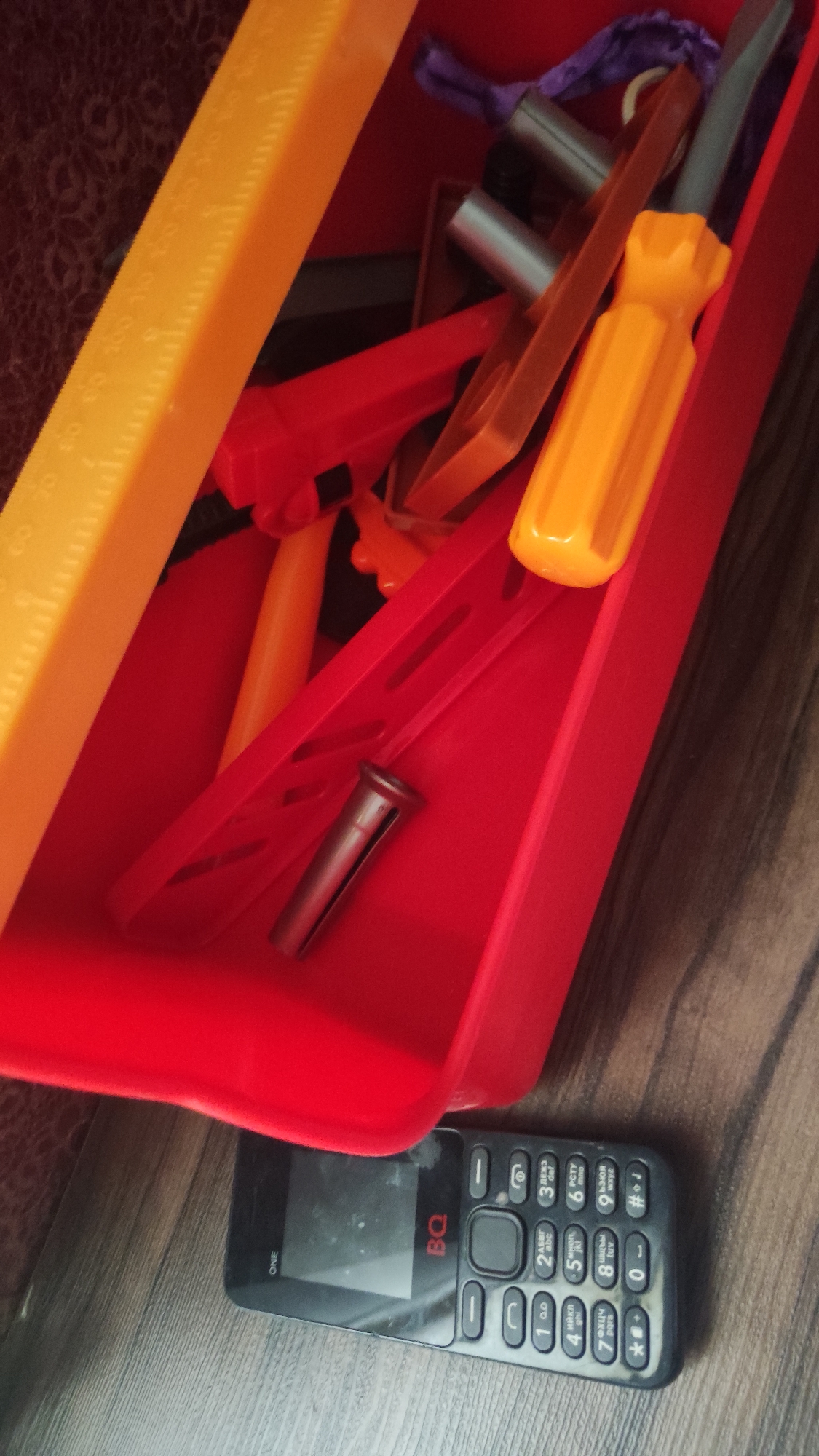 A red plastic toolbox containing various children's plastic tools, including a yellow ruler, a yellow screwdriver, and other red and orange items, sits on a wooden surface. A black BQ-branded feature phone rests next to the toolbox. The photograph appears to be taken indoors. The location within Chaikovsky, Russia, cannot be determined from the image alone.