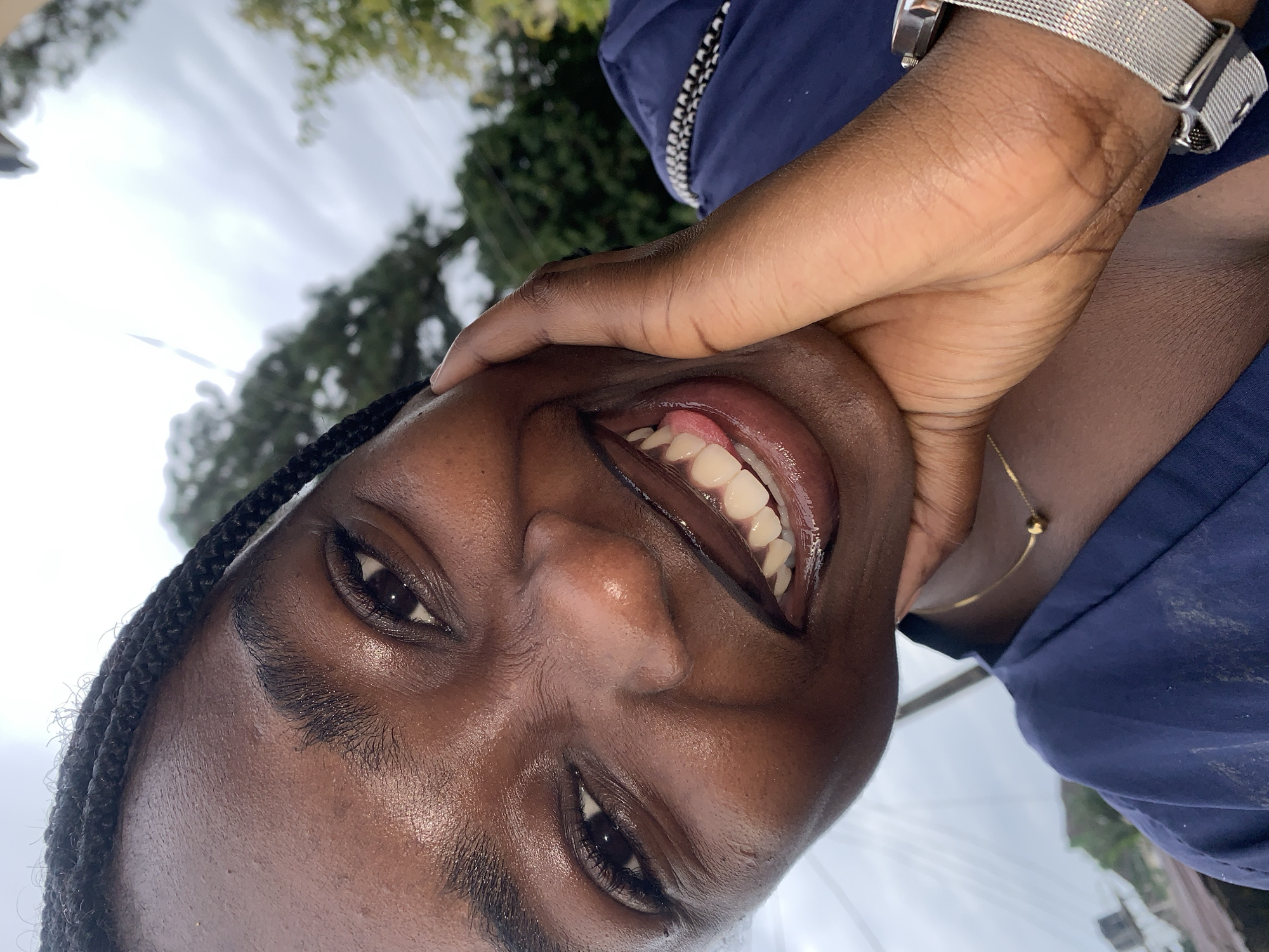 A close-up, eye-level outdoor portrait shows a young Black woman smiling widely, her right hand resting on her chin, with her tongue peeking out. The woman has braided hair pulled back, and her skin is shiny, suggesting it may be dewy or oily. She is wearing a dark blue V-neck garment and a thin gold necklace with a small bead pendant. A silver watch with a mesh band is visible on her right wrist. The background is blurred, showing greenery and a cloudy sky. The image is cropped to focus on the woman's face and upper body. The location is identified as Rafin Par, Nigeria.