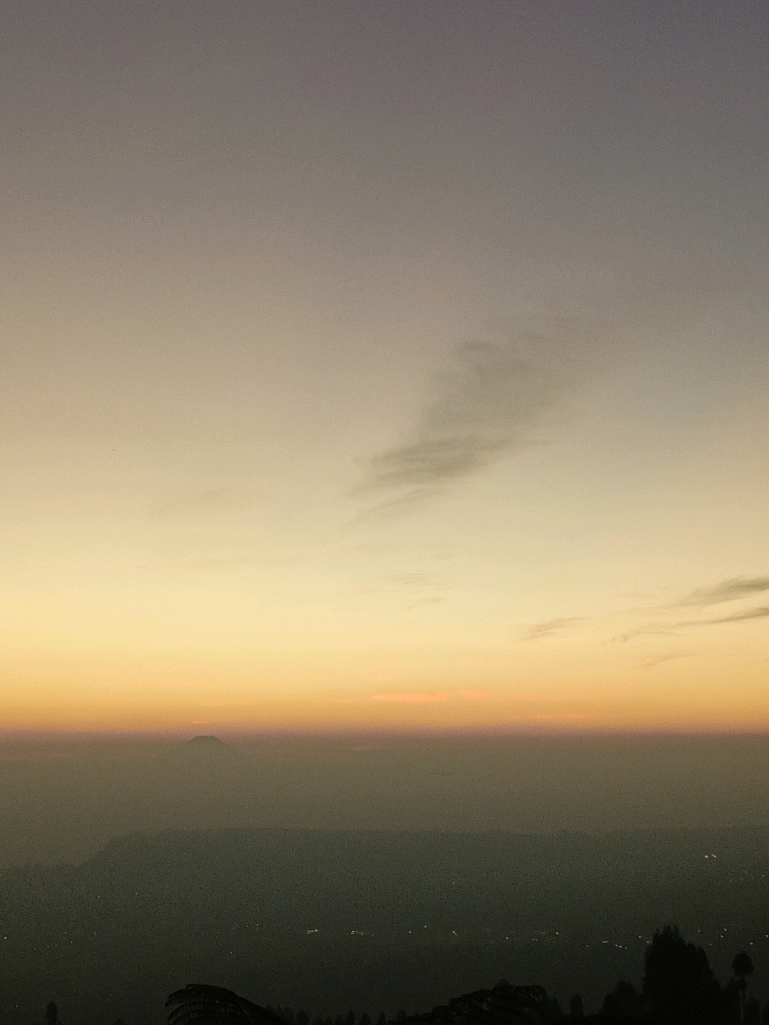 This is a horizontal, medium shot photograph taken at dusk, overlooking a valley from a high vantage point in or near Butuh Kidul, Indonesia. The sky dominates the upper two-thirds of the frame, transitioning from a pale lavender at the top to a warm, gradient orange and yellow band just above the horizon. Faint, wispy clouds are scattered across the upper sky. The lower third of the image depicts a hazy, dark green valley filled with the faint lights of a settlement. In the distance, a silhouetted mountain with a flattened top rises above the valley mist. In the foreground, the dark silhouettes of trees and what appears to be a large, fan-like structure are visible at the bottom edge of the frame. The overall atmosphere is serene and expansive, capturing the transition from day to night over a rural or semi-rural Indonesian landscape.