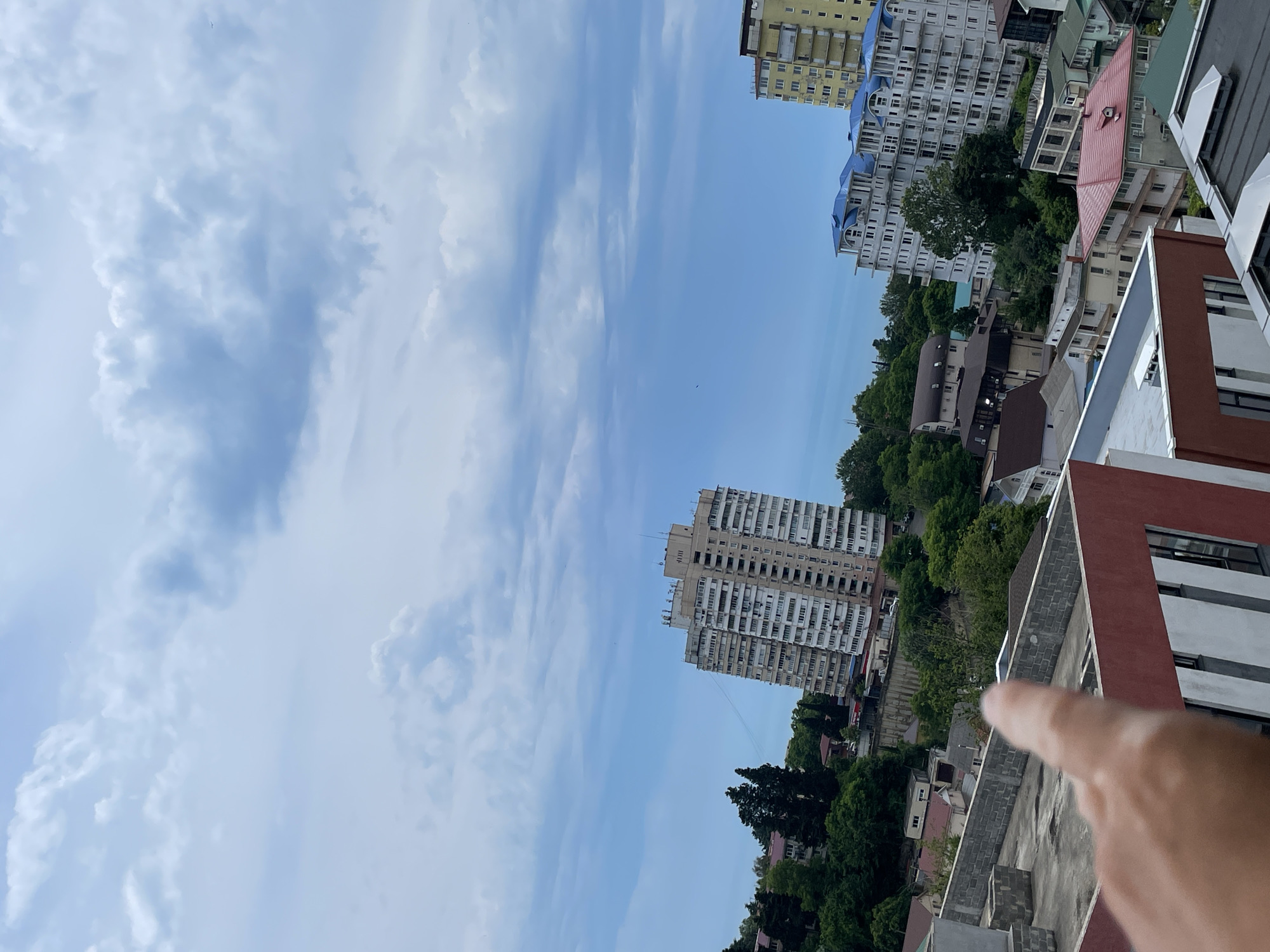 This is an outdoor, eye-level, vertical, medium shot of a cityscape in Sochi, Russia. The image is taken from a high vantage point, looking down and out over buildings and greenery. The sky above is filled with dramatic, grey and white clouds against a pale blue backdrop. In the mid-ground, a tall, beige apartment building with many windows is visible, leaning slightly to the right. To the right of that building, a taller, yellow building with balconies stands out. In front of these, and closer to the viewer, are several white apartment buildings with distinctive blue roofs, some appearing to be under construction. Below these structures, a dense canopy of green trees covers rolling hills, interspersed with smaller residential buildings. In the foreground, a hand with an extended index finger, pointing upwards and to the right, is visible from the elbow down. The hand is in focus, obscuring a portion of the rooftop in front of it. The rooftop appears to be made of concrete and has grey brick walls.