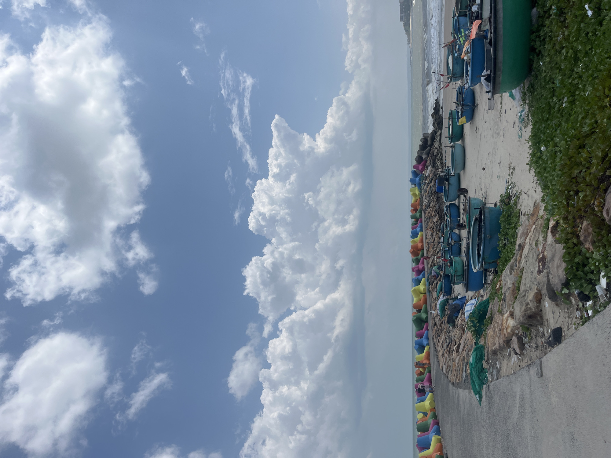 The image depicts a coastal scene in Phú Thủy, Vietnam, on a bright, partly cloudy day. A concrete pathway lines the left side of the frame, leading towards a colorful barrier of tetrapods (concrete breakwater blocks) in shades of blue, green, yellow, and orange. Beyond the tetrapods, a sandy beach is visible, cluttered with numerous blue and green basket boats, some appearing empty and others with fishing gear. The sea, with moderate waves, stretches towards the horizon, where a town or resort is faintly visible on a distant headland. The foreground shows scattered rocks and low-lying green vegetation.