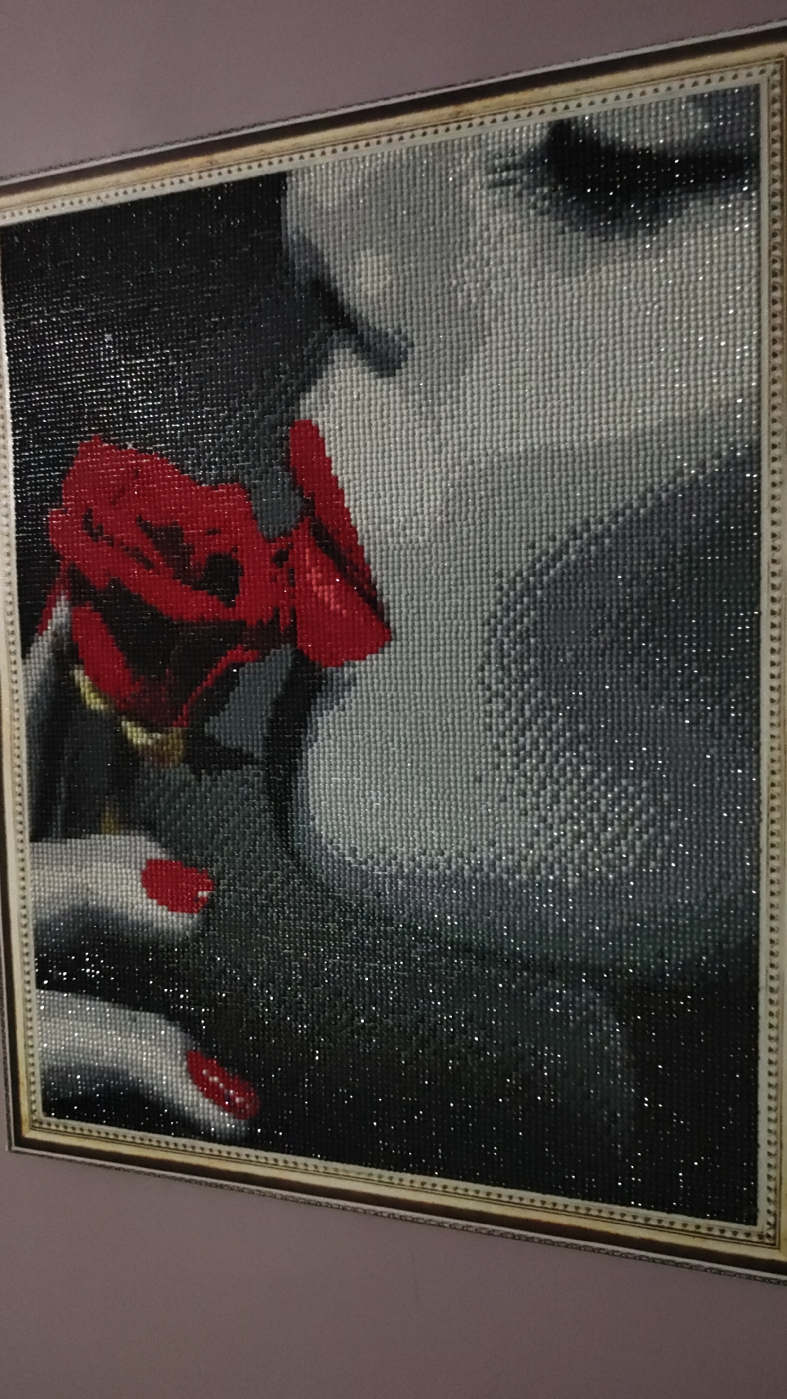 This media file features a framed diamond painting, showcasing a close-up profile of a woman with a red rose. The artwork predominantly uses a monochrome palette of black, white, and various shades of grey, with the vibrant red of the rose and the woman's nail polish providing strong focal points of color.
The main subject is a woman whose face is partially visible from her nose to her chin, with her lips delicately parted. Her hand, adorned with striking red nail polish, gently holds the stem of a lush red rose, bringing it close to her face. The pose evokes a sense of intimacy or contemplation, as if she is inhaling its scent or simply appreciating its beauty. The pixelated texture characteristic of diamond paintings is clear, with thousands of small, sparkling resin beads forming the image and creating a shimmering effect across the surface, particularly in the darker areas and highlights of her hair and attire.
The painting is encased in a decorative, light-colored frame, possibly golden or silver, which provides a classic border to the modern medium. This framed piece is displayed on a wall painted in a soft, muted purple hue. The overall impression is one of elegance and romance. There is no visible text within the image.