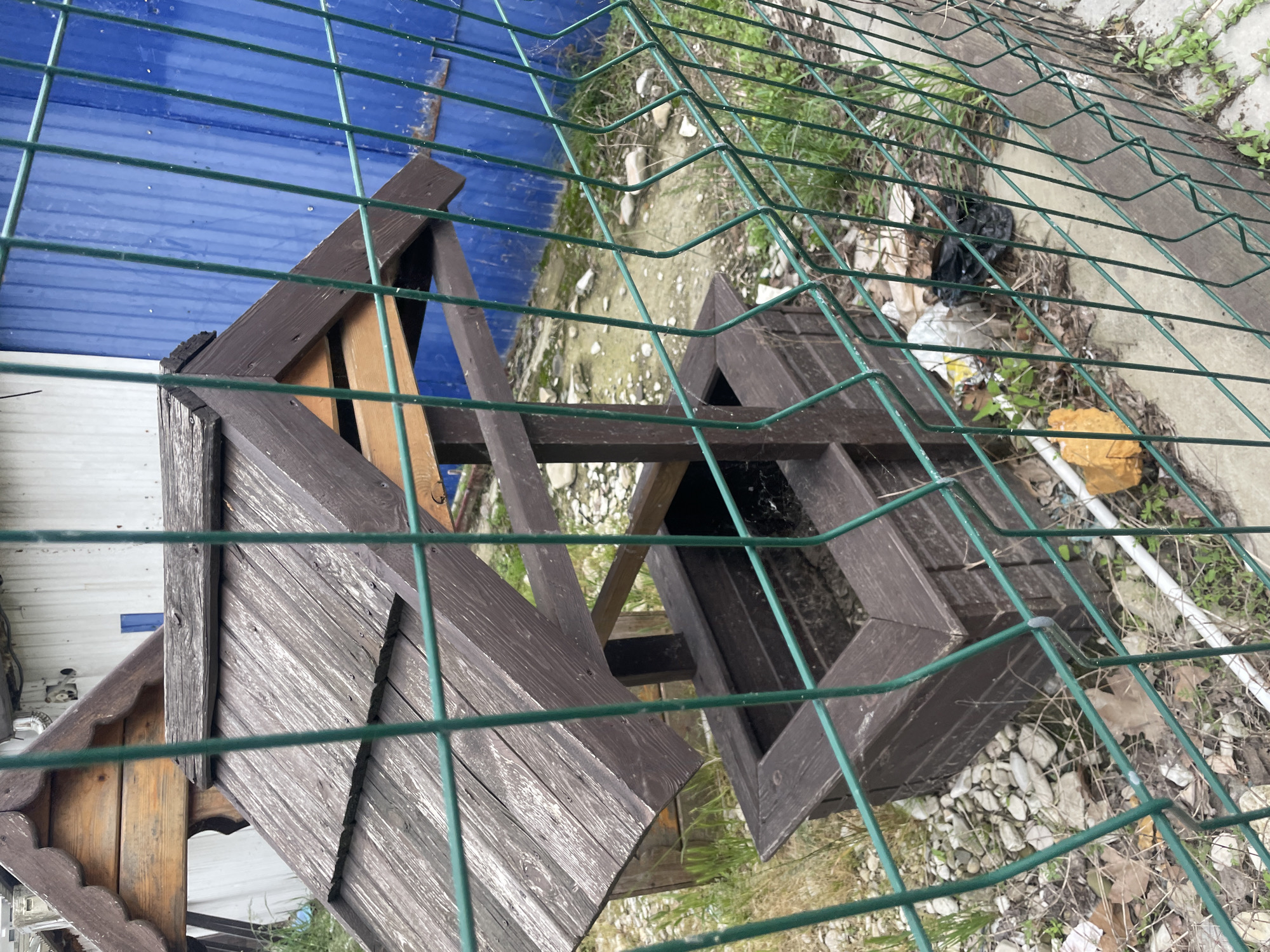 A wooden birdhouse and planter stand behind a green chain-link fence. The planter is empty and appears to be in disrepair. The birdhouse has a shingled roof and decorative trim. The ground behind the fence is covered with rocks, leaves, and debris. In the background are glimpses of a blue corrugated metal structure and other buildings. The location is presumably Olginka, Russia, based on the user's prompt.