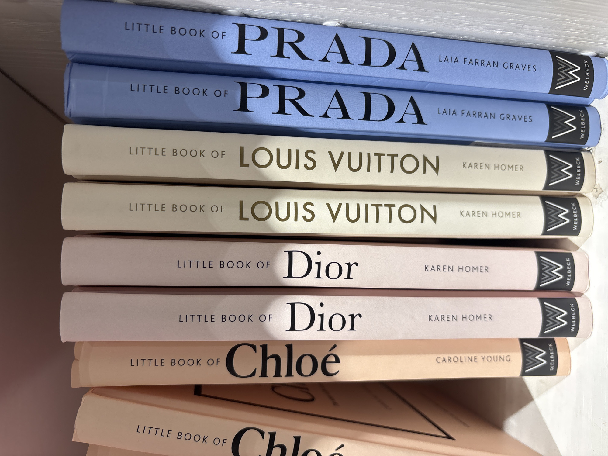 The image shows a stack of books, specifically the "Little Book of..." series by Welbeck Publishing. From top to bottom, the books are "Little Book of Prada" by Laia Farran Graves (two copies), "Little Book of Louis Vuitton" by Karen Homer (two copies), "Little Book of Dior" by Karen Homer (two copies), and "Little Book of Chloé" by Caroline Young (two copies). The Prada books are a light blue, the Louis Vuitton books are off-white, the Dior books are light pink, and the Chloé books are light peach. The scene is not located in the city of Sofia, Bulgaria. It shows a shelf of books.