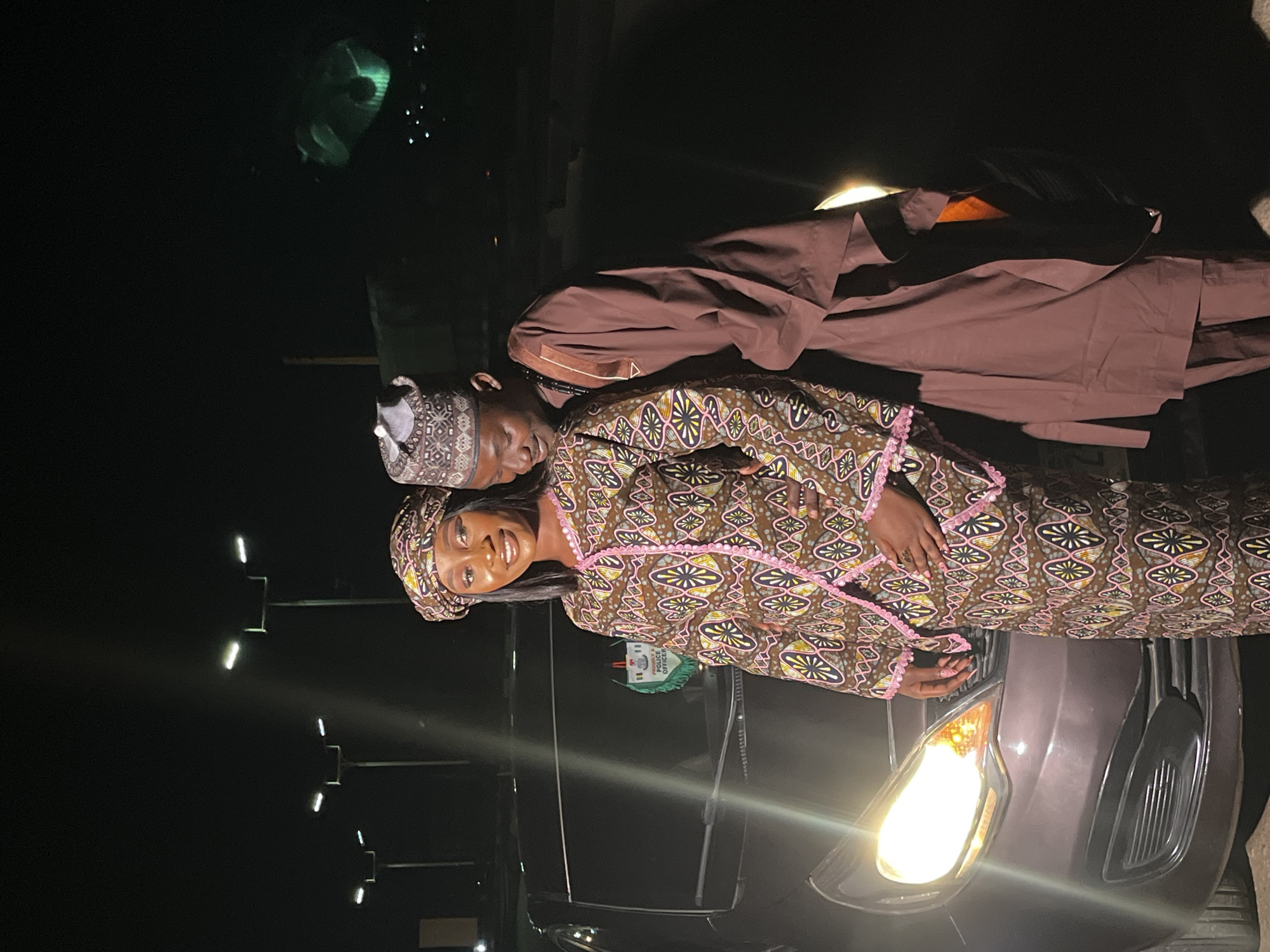 A couple is posing in front of a car at night in Gura Top, Nigeria. The woman is wearing a patterned traditional outfit with a matching headscarf, and the man is wearing a brown traditional outfit and cap. They are standing close together, with the man's arm around the woman. The car's headlights are on, illuminating the scene. Streetlights are visible in the background.