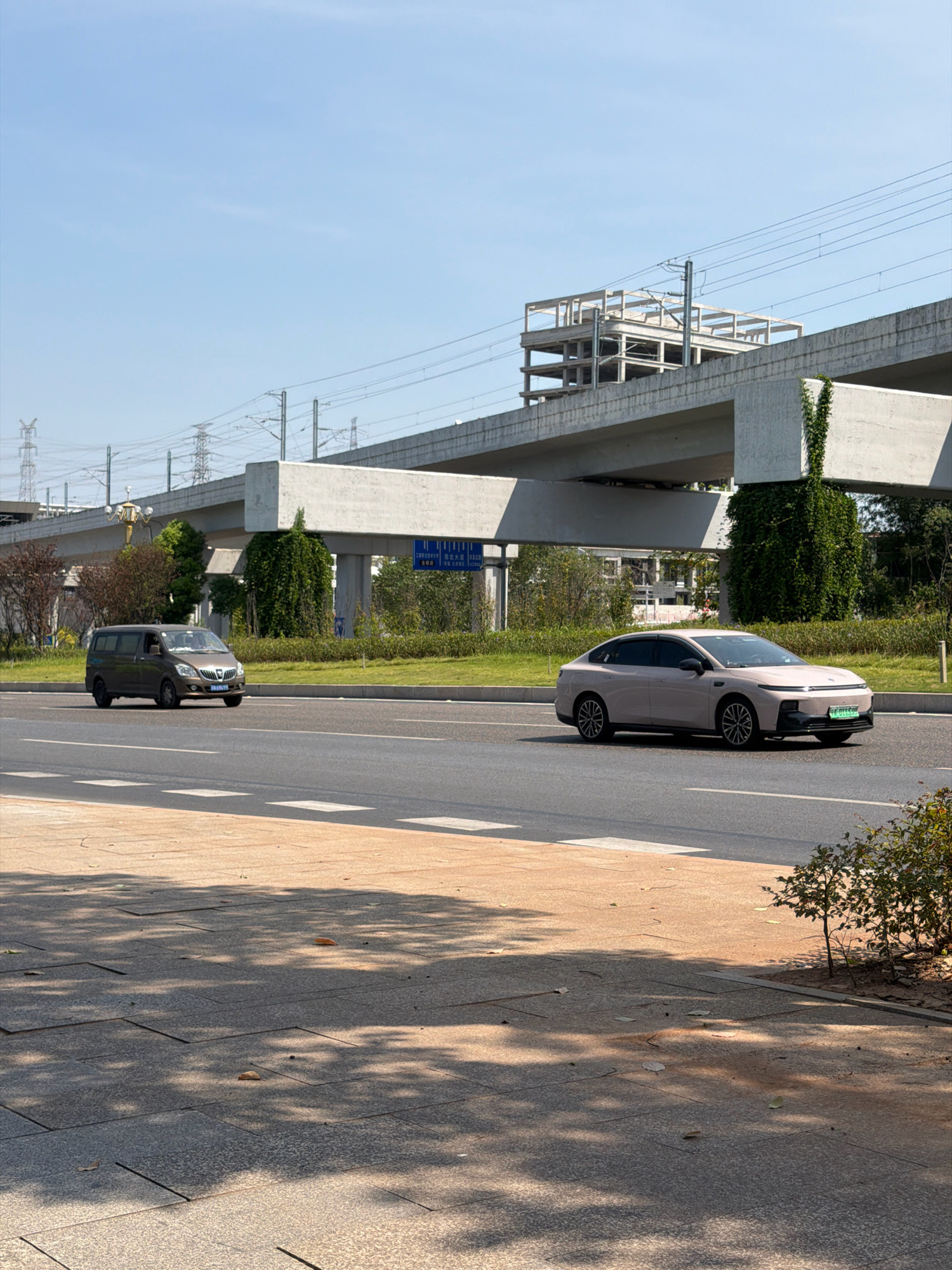 The image depicts a street scene in the city of Jiangjin, China, with a focus on vehicular traffic and elevated infrastructure. A light pink sedan is traveling on the right side of the road, while a brown minivan is on the left. Overhead, a concrete elevated structure, possibly a highway or train line, spans across the scene, supported by pillars. Construction elements, such as rebar, are visible on top of this structure, suggesting ongoing development. Power lines and utility poles are also present in the background, indicating an urban or semi-urban environment. The sky is clear and blue, and the sun casts shadows on the paved pedestrian area in the foreground. Lush green vegetation lines the sides of the road and the base of the elevated structure, suggesting a well-maintained urban landscape. The overall atmosphere is one of a functional, developing city with active transportation routes.