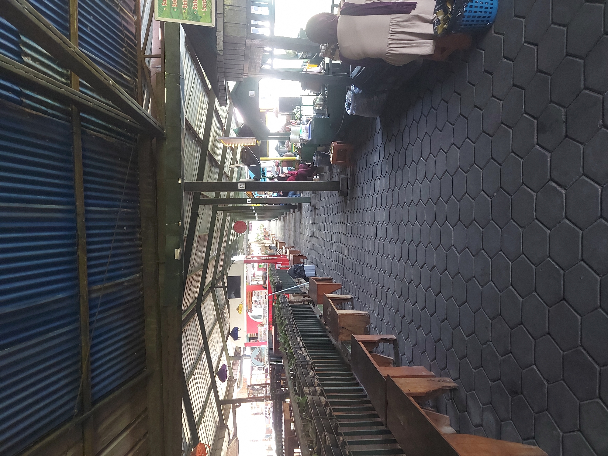 The image depicts an outdoor covered walkway, likely part of a market or food court in Lembang, Indonesia. The walkway is paved with dark hexagonal tiles and lined on the left with wooden benches and planters filled with greenery. On the right, several food stalls are visible under a corrugated metal roof supported by wooden posts. The stalls are open-fronted, showcasing food preparation areas and seating.
Several people are present in the scene. On the left side, a woman wearing a hijab is seated at a stall. Further down the walkway, more individuals can be seen in the background, some sitting and others standing. On the right side, a woman wearing a light-colored dress and a purple hijab is standing near a blue trash bin.
The environment appears to be a public space designed for casual dining and shopping. The architecture suggests a rustic and functional design, with a weathered metal roof and simple wooden structures. The lighting indicates it is daytime, with sunlight filtering through the roof and casting shadows. Decorations, such as colorful hanging lanterns, add a festive touch. Signs are visible on some stalls, one appearing to list food items. The overall impression is of a lively, yet somewhat weathered, local market.