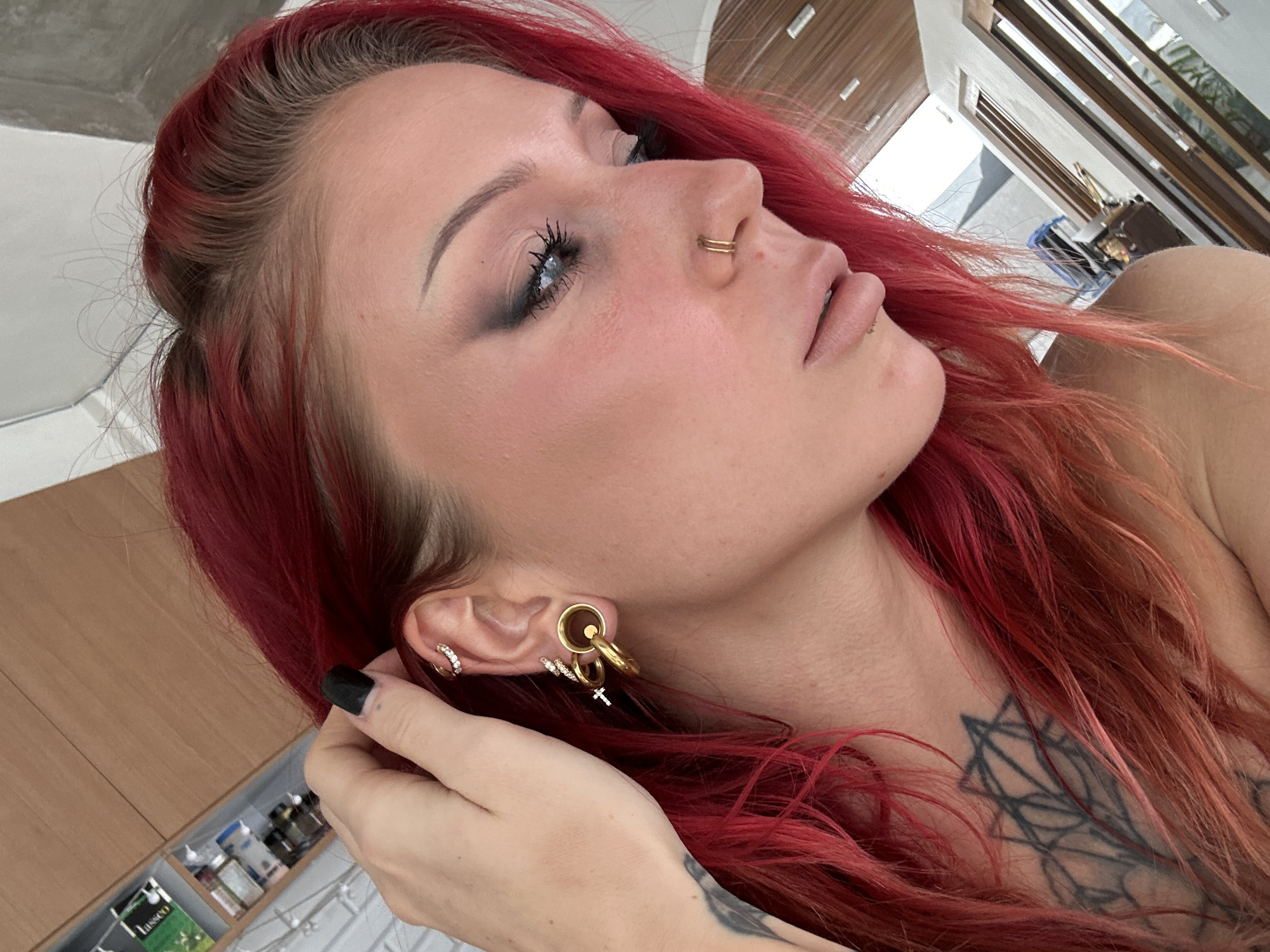A woman with bright red hair is shown in a close-up, profile view. She is wearing large gold hoop earrings, a smaller hoop earring with diamonds, and a cross charm earring in her left ear. She also has a double hoop nose ring. The background appears to be an indoor setting with wooden paneling and large windows, possibly in a modern residence or resort. There are some shelves with items visible on the left side of the image. The woman has dark eye makeup and is looking down and to the right. A tattoo is visible on her chest. The lighting is natural, suggesting it might be daytime. The location is identified as North Kuta, Indonesia.
