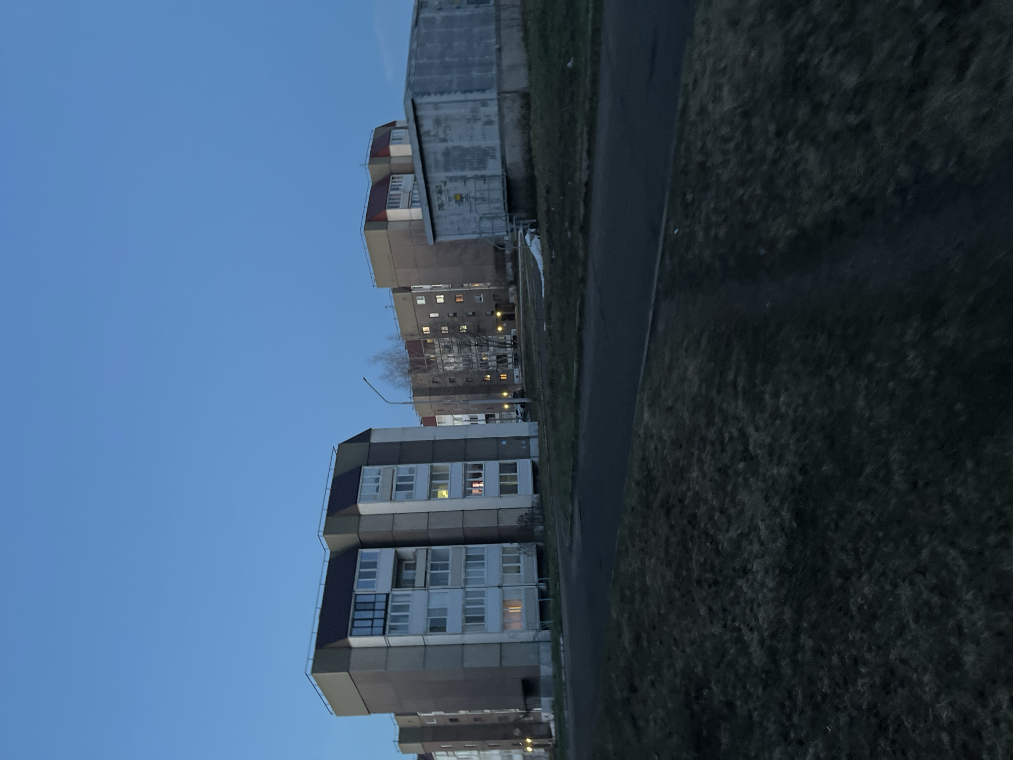The image depicts an urban scene in Марковский, Russia, possibly taken during the evening or early morning, indicated by the blue sky and some illuminated windows in the buildings. The buildings appear to be typical Soviet-era apartment blocks, arranged in a row. A paved road or pathway runs along the side of the buildings, bordered by grassy areas. The image is oriented vertically, showing the buildings at an angle.