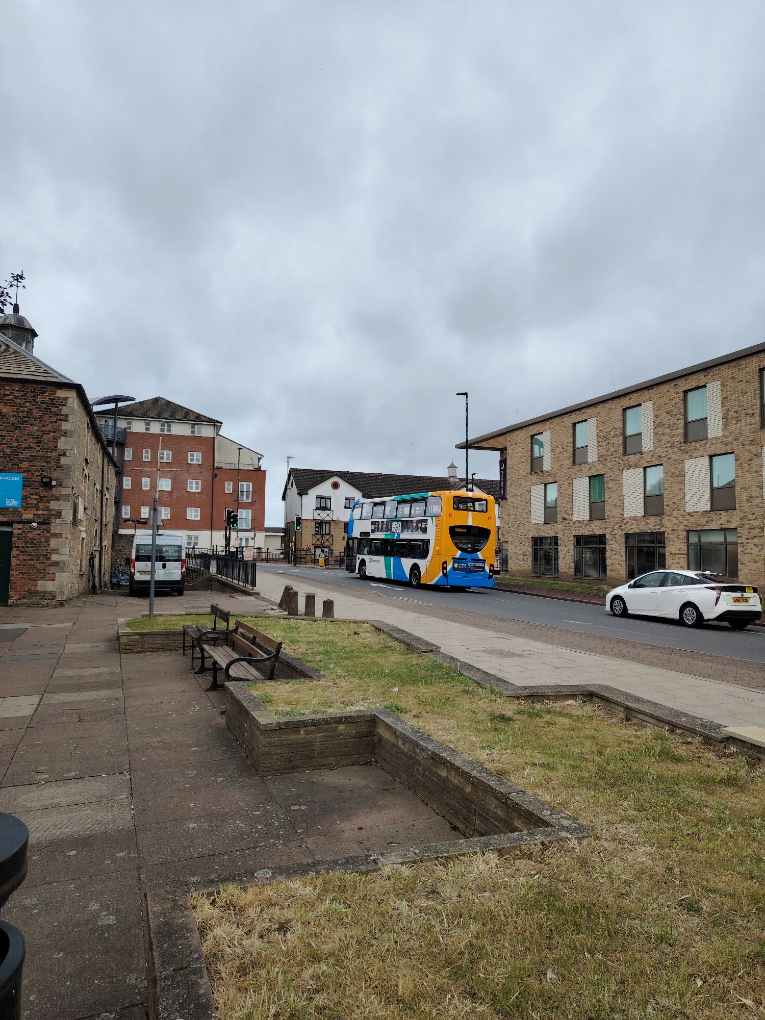The image presents an urban street scene in Peterborough, United Kingdom, viewed on an overcast day.
In the immediate foreground, a grey paved sidewalk extends from the lower left to the upper right. On the left side of this sidewalk, two dark brown wooden benches with black metal frames are positioned. To their right, two rectangular concrete planters contain dry, light brown and green grass. A dark circular bin is partially visible in the lower-left corner.
A strip of dry, unkempt grass separates the sidewalk from a two-lane asphalt road.
On the left side of the road, a building constructed from light brown stone features a dark tiled roof and a small cupola topped with a weather vane. A blue sign affixed to its facade clearly reads "RNLI PETERBOROUGH CHARITY SHOP." A white panel van, displaying some green text, is parked adjacent to this building, slightly recessed. A traffic light showing a green signal is visible near the van.
On the road, a yellow, blue, and white Stagecoach double-decker bus, featuring an advertisement for "SCARY MOVIE," is seen driving away from the viewer. Behind and to the right of the bus, a white Toyota Prius (registration ENE7 HZG) is also traveling in the same direction.
On the right side of the road, a modern, multi-story building constructed from light brown brick features numerous windows with dark frames and some white brick sections. A subtle purple sign indicating "Premier Inn" is partially visible on the building's side.
In the background, further down the street, a taller brick building with multiple white-framed windows and balconies is visible on the left. To its right, a smaller white building with dark timber detailing is partially obscured.
The sky is uniformly grey with dense cloud cover. No individuals are discernible within the scene.
