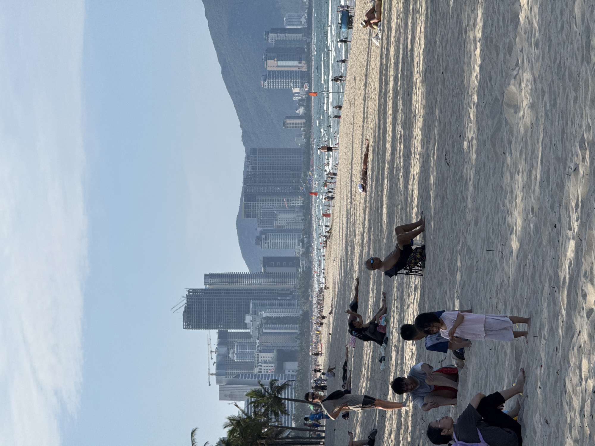 The image shows a crowded beach in Đà Nẵng, Vietnam. A long stretch of sandy beach is filled with people. Many are lying down on the sand or standing near the water. Palm trees line the back of the beach, with tall buildings visible in the background. Some buildings appear to be under construction. A mountain is visible in the distance on the left side of the image. The sky is blue with some clouds. Shadows from palm trees cast dark stripes on the beach. Several small children are on the beach. An older man in a wheelchair is on the beach.