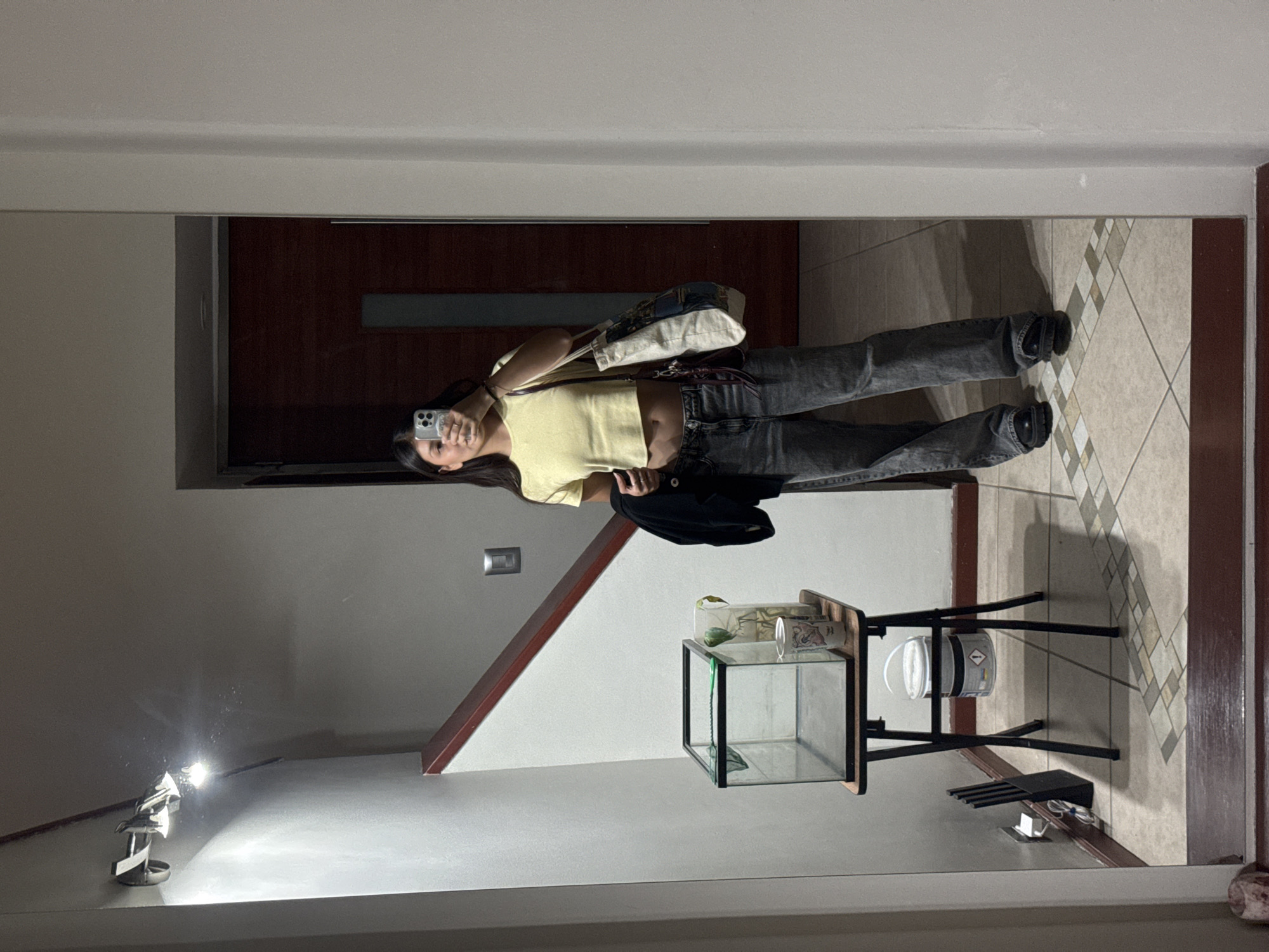 The image depicts a person taking a mirror selfie in an indoor environment in Heroica Puebla de Zaragoza, Mexico. The person is wearing a yellow crop top, grey jeans, and black shoes. They are holding a cell phone in their right hand to take the picture. A bag hangs from their left shoulder. The background features a hallway with a dark wooden door, a staircase with a red-brown railing, and a tile floor. A glass terrarium stands on a black metal stand next to a wall. A white cylindrical object sits on the lower shelf of the stand. A light fixture is visible at the bottom left. An electrical outlet is mounted on the wall next to the stairs.
