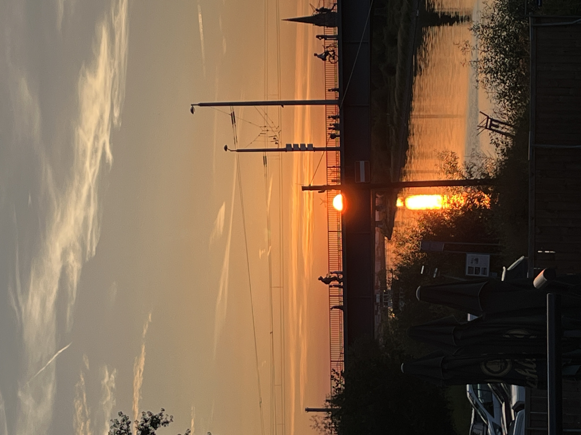 The image captures a sunset over a body of water, likely a river, in an urban environment. A silhouetted bridge spans the water, with several figures visible walking and cycling across it. The sun is low on the horizon, casting a warm, orange glow across the sky and reflecting brightly on the water's surface. There are streetlights and power lines visible on and around the bridge, indicating a developed area. In the foreground, partially closed umbrellas suggest an outdoor seating area, possibly a cafe or restaurant, overlooking the scene. The overall impression is one of a serene evening in a city.