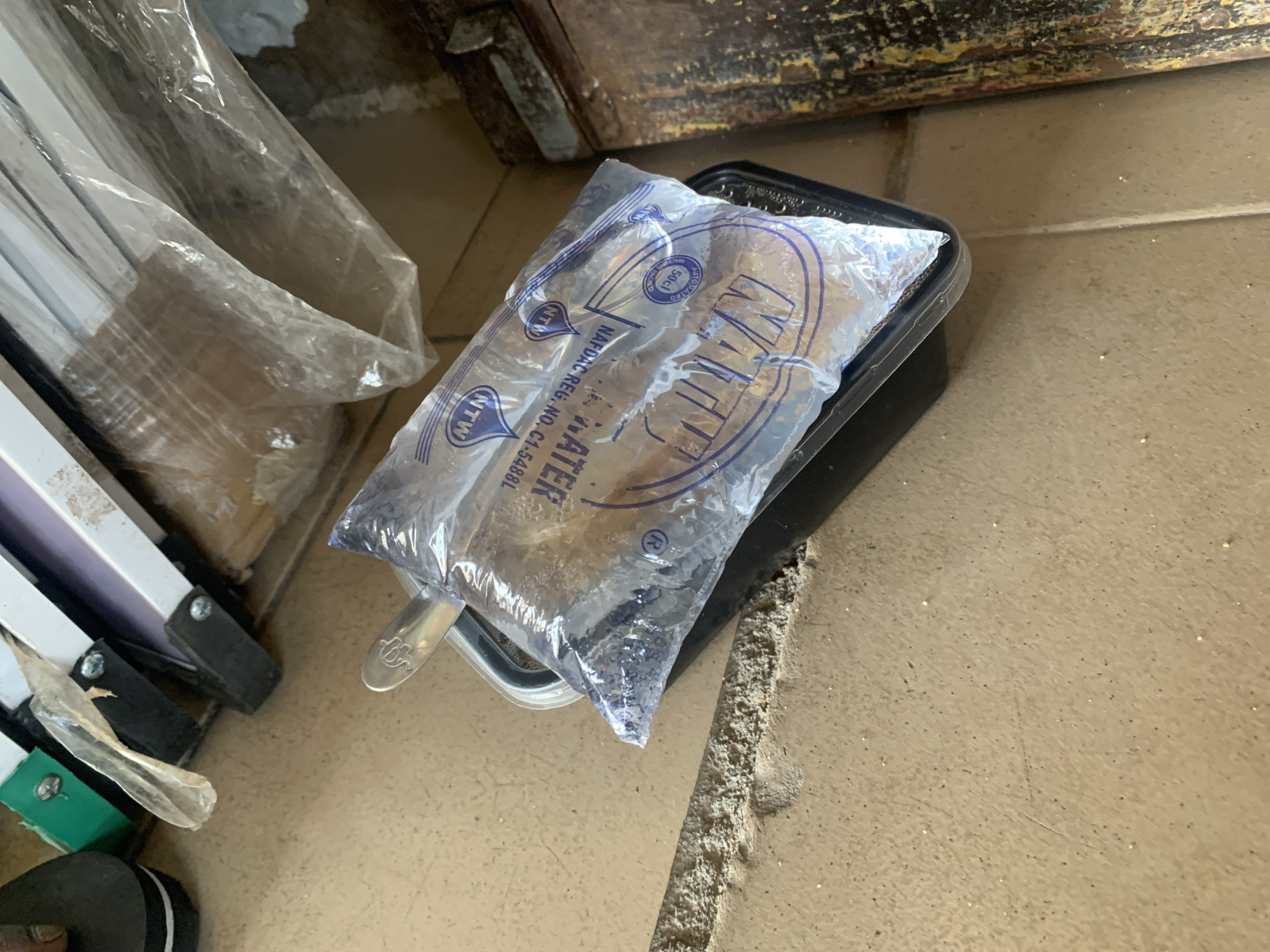 A plastic bag of water, branded "NTN," rests on a black plastic container. The bag appears to have condensation on its surface. The container is placed on a tan-colored, tiled floor. In the background, a portion of a white metal structure with some plastic sheeting is visible. To the left of the frame, a dark-colored sandal or shoe is partially visible. To the right, a rusty, weathered metal object is present. The lighting suggests an outdoor or well-lit indoor environment. There are no people visible in the image.
